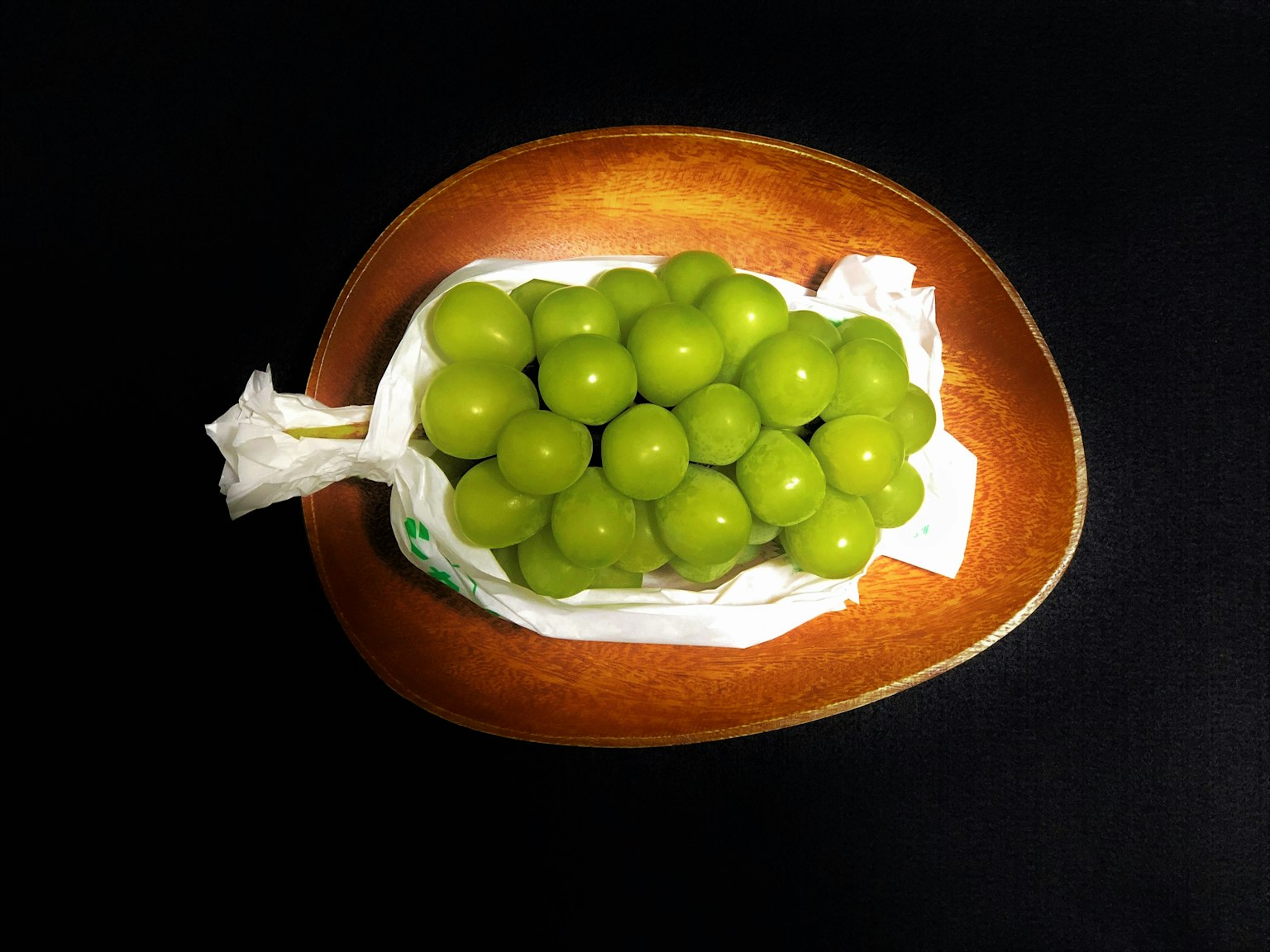 องุ่นเขียวในถุงสีขาวบนจานไม้พื้นหลังสีดำ