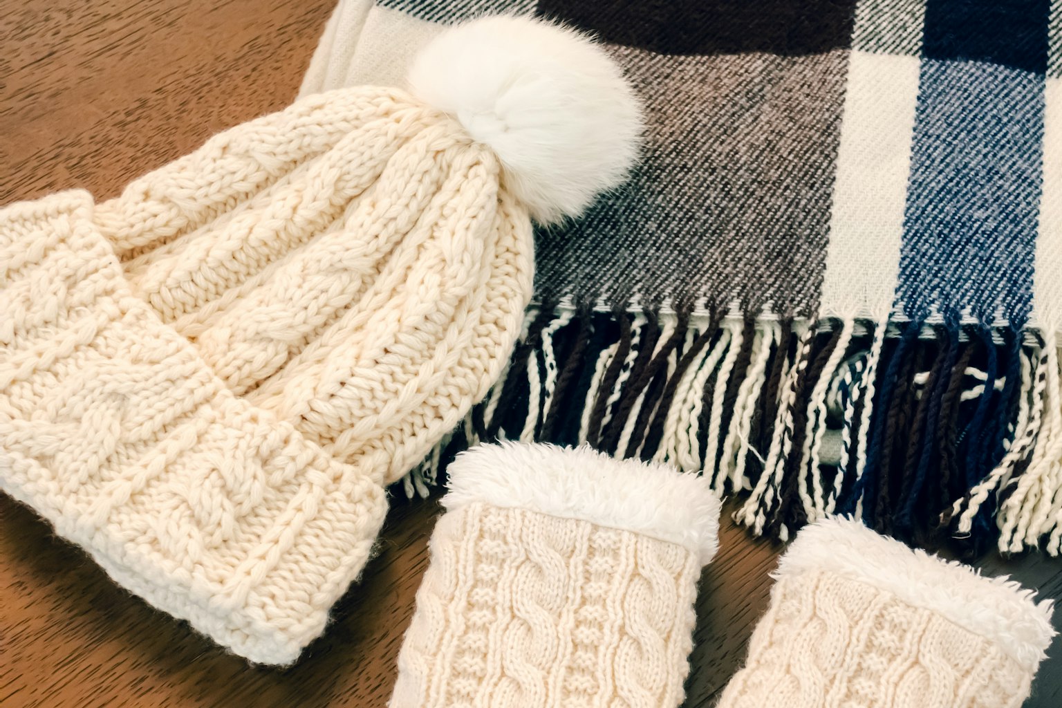 Cremfarbener Strickmütze mit Pompon und passende Handschuhe neben einem karierten Schal