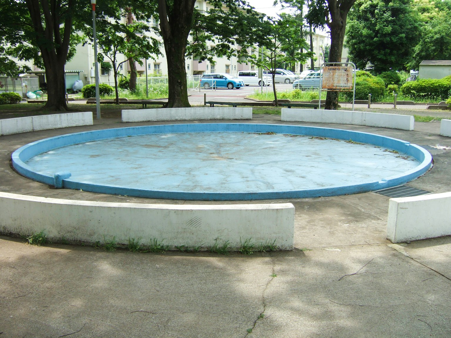 公園內的圓形藍色游泳池，周圍有白色長椅和樹木