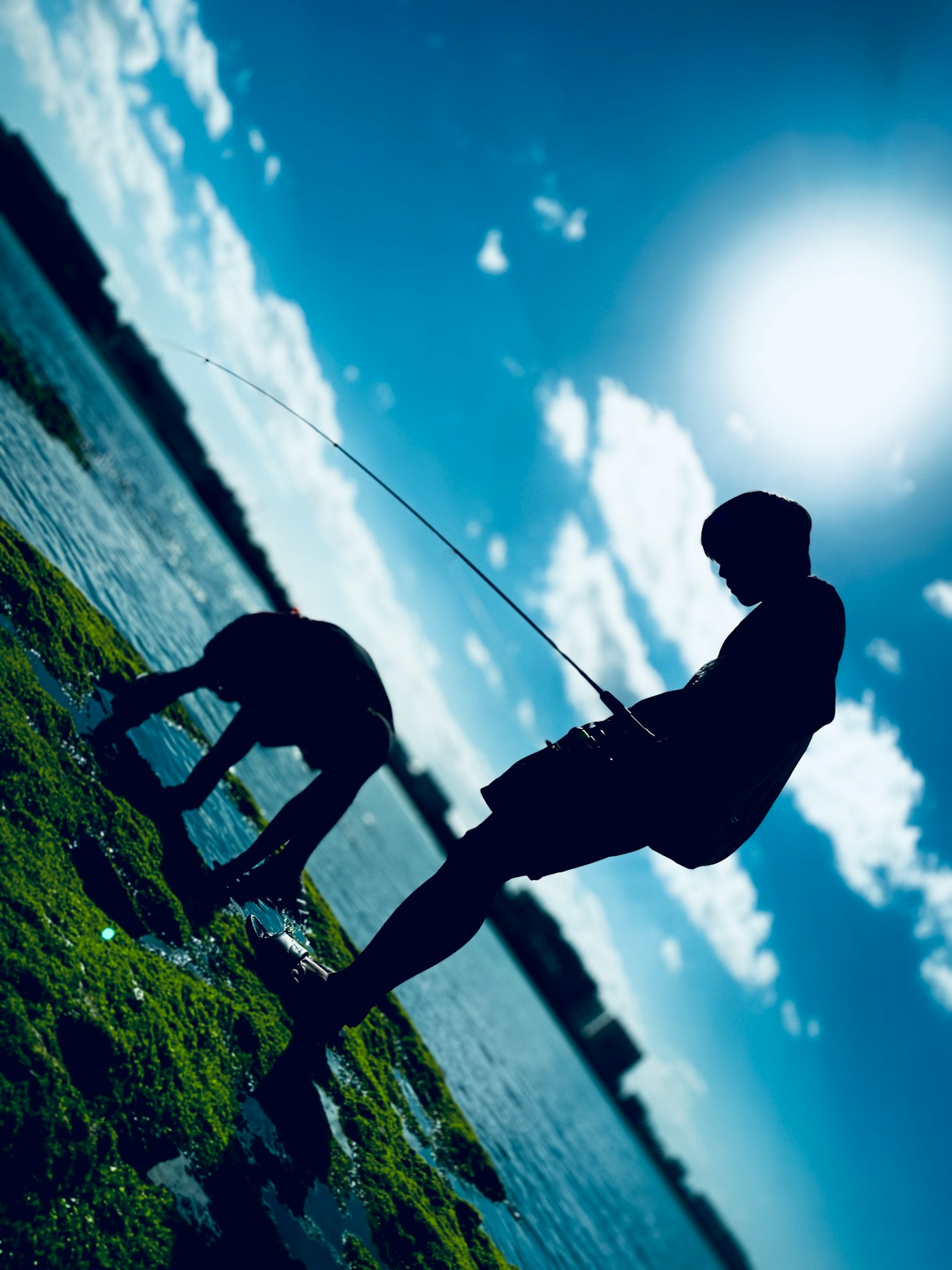 青い空の下で釣りをする二人のシルエットと太陽