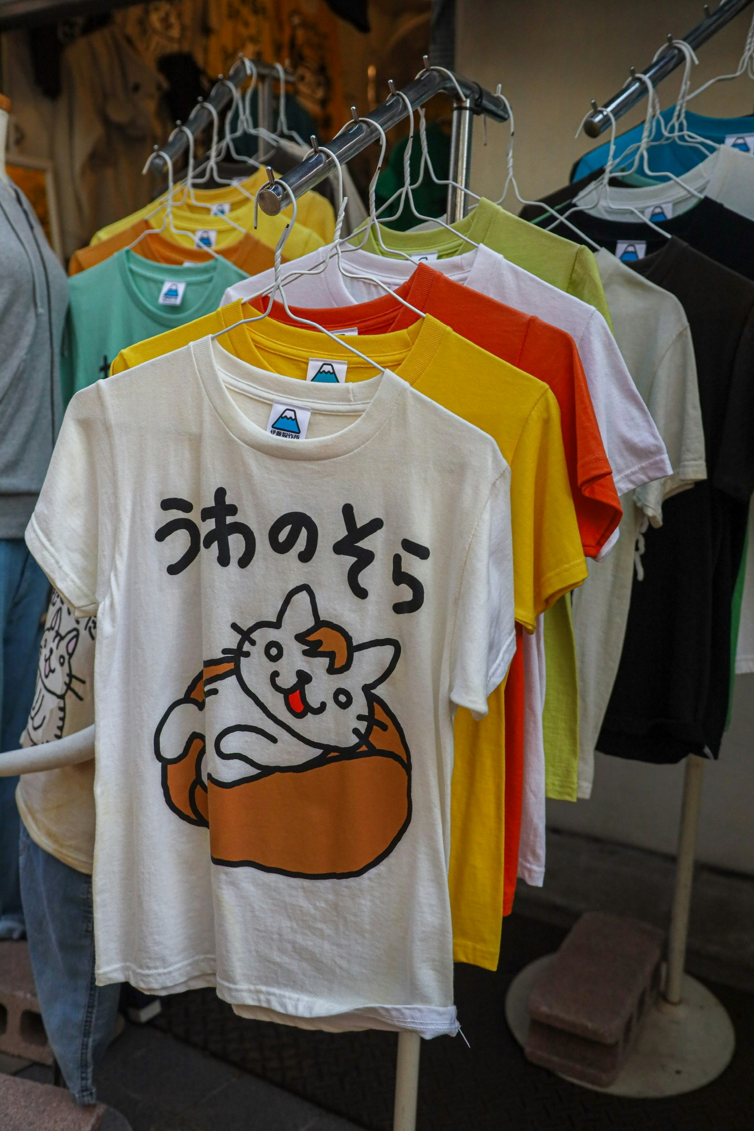 เสื้อยืดหลากสีแขวนอยู่บนไม้แขวนเสื้อโดยเสื้อยืดขาวมีภาพวาดแมวและข้อความภาษาญี่ปุ่น