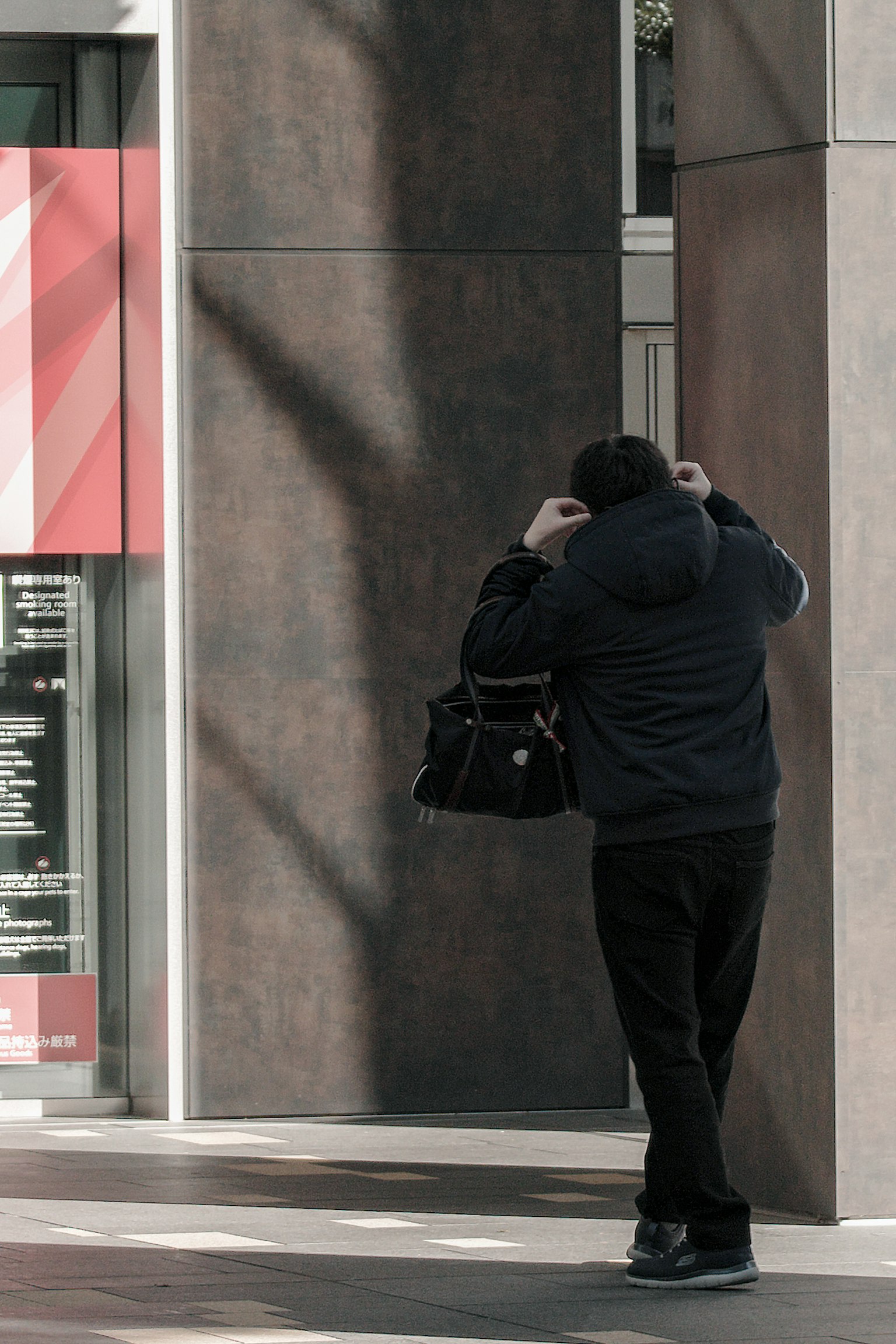 Person standing with back to wall wearing dark outerwear holding a bag subtle shadows in urban setting