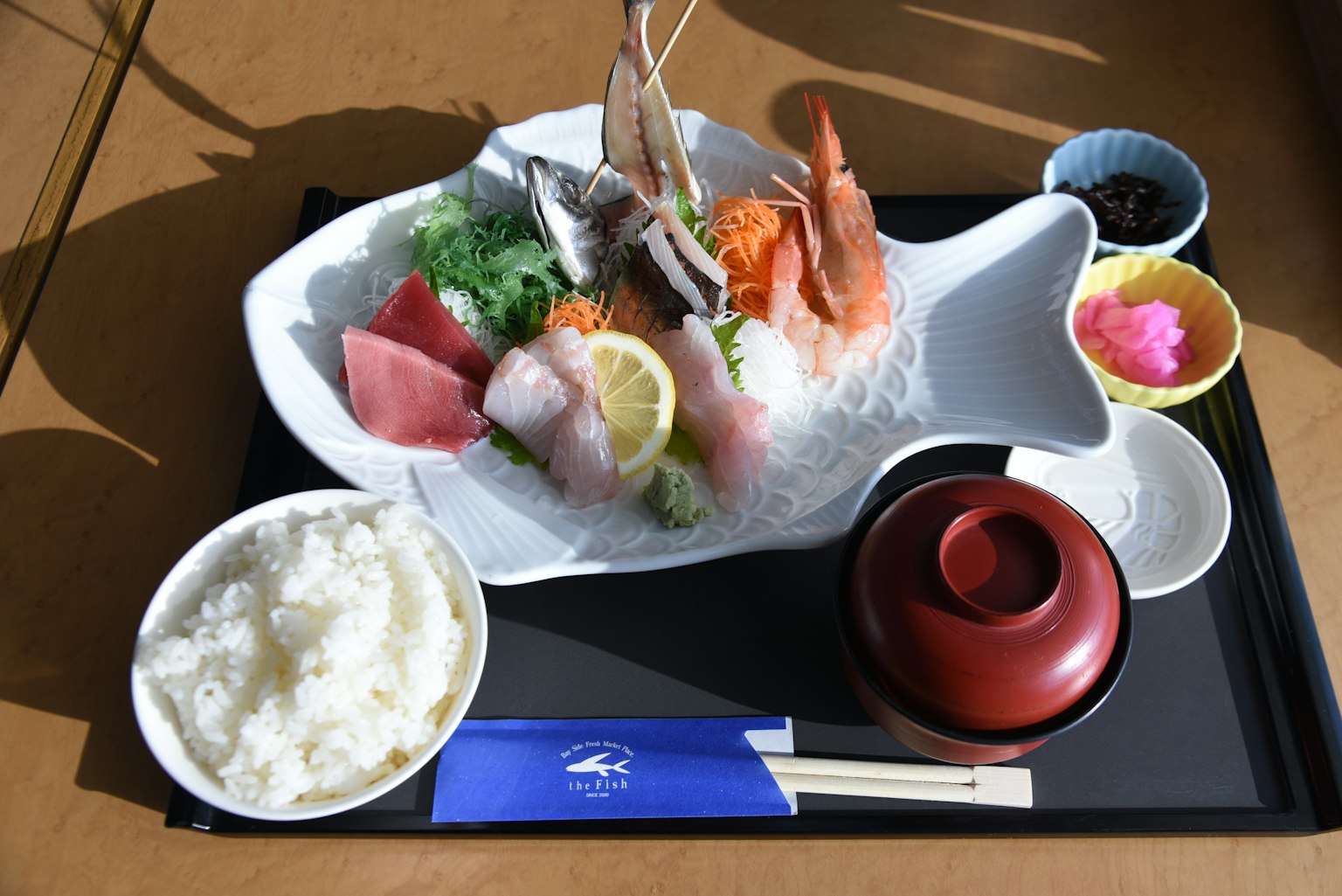 Piring yang menampilkan berbagai sashimi segar disajikan di atas piring berbentuk ikan dengan nasi dan sup miso