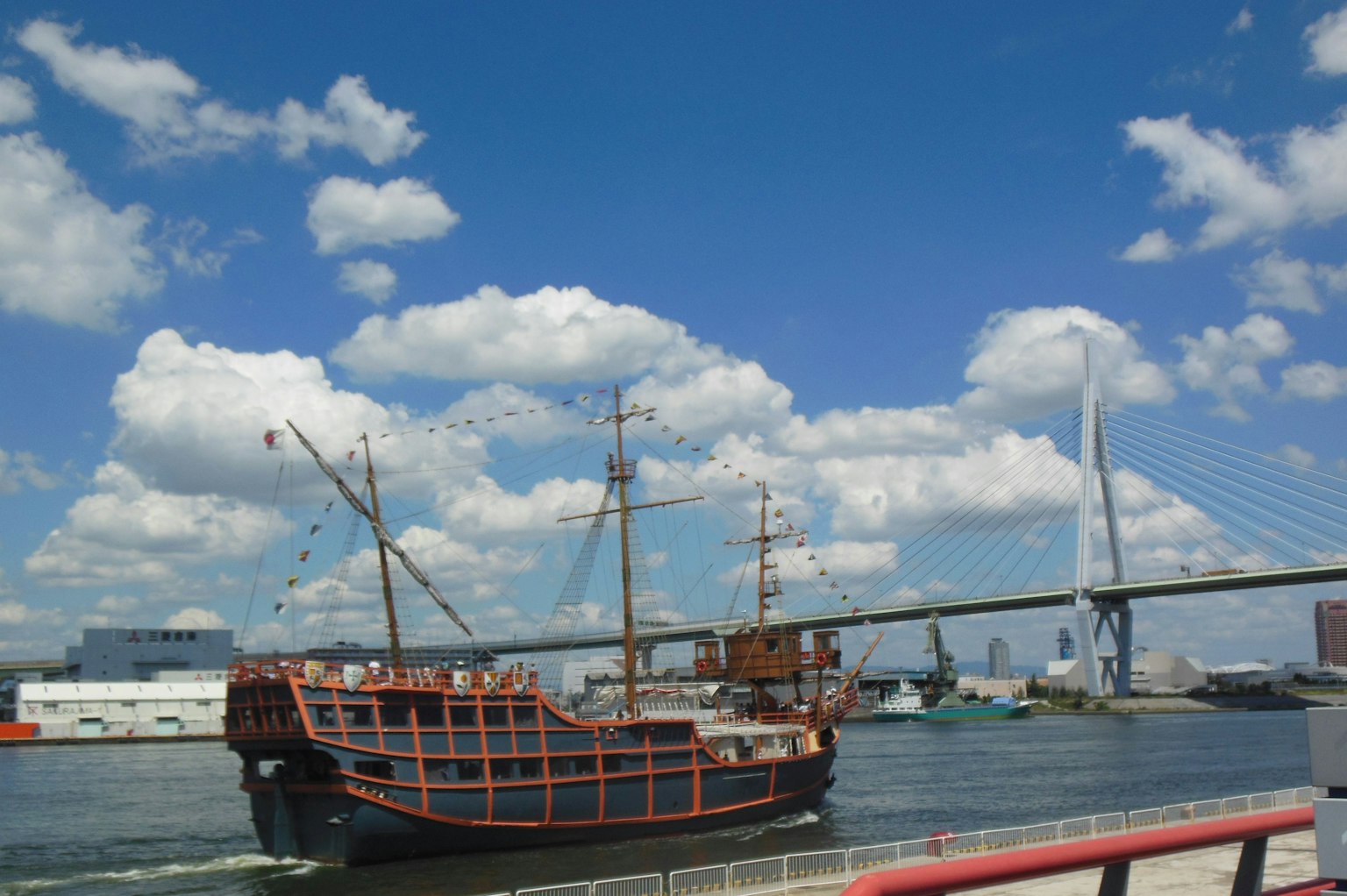 Kapal layar tradisional di bawah langit biru dengan awan dan jembatan modern