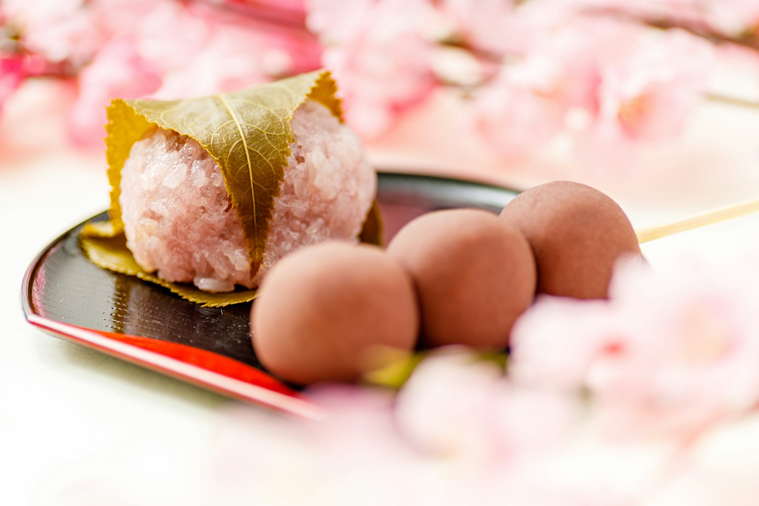 Presentazione di dolci giapponesi circondata da fiori di ciliegio con mochi sakura avvolto in foglie e tre dango