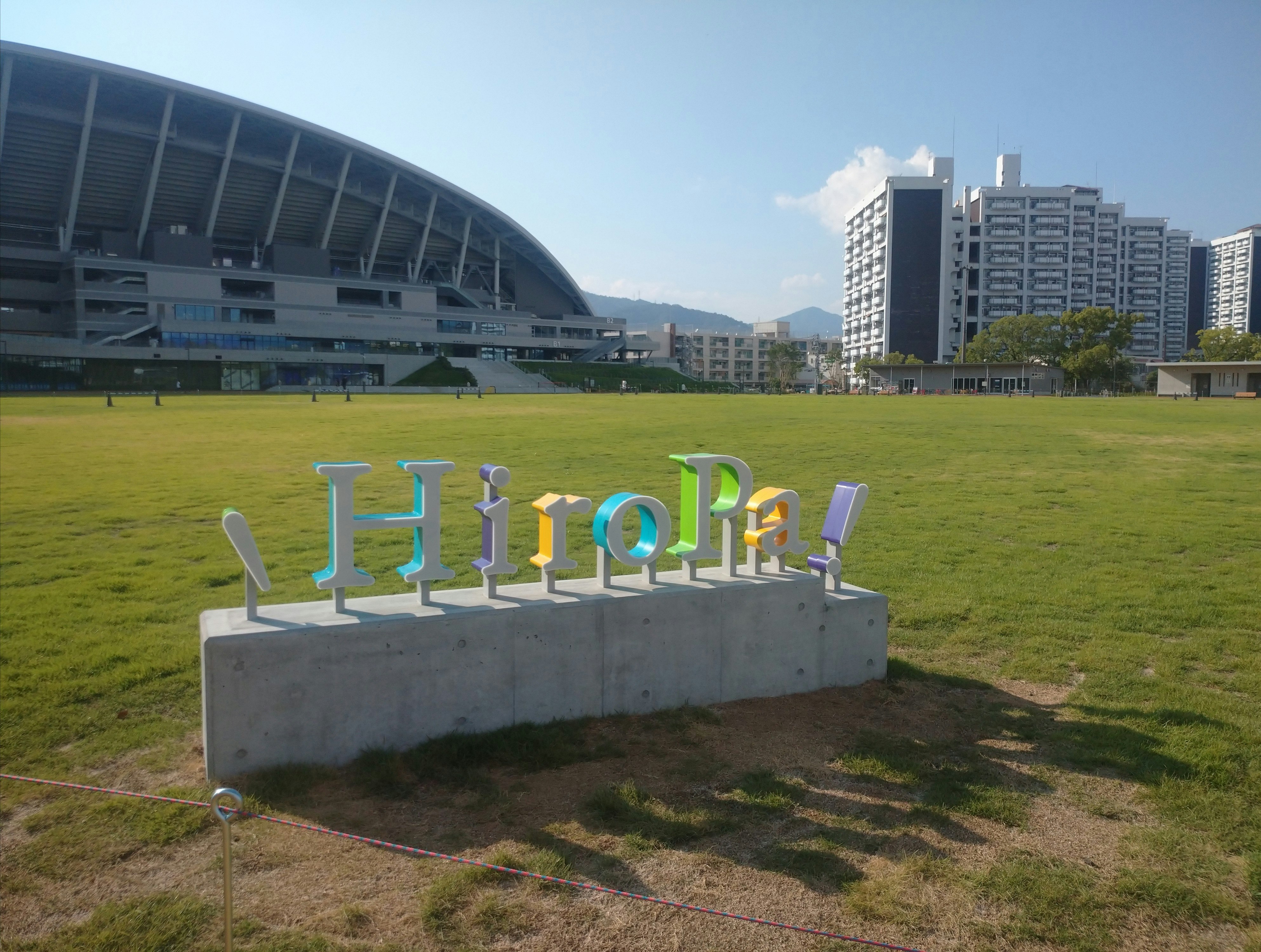 芝生の公園と「Hello Pal」モニュメント、スタジアム風の建物が見える