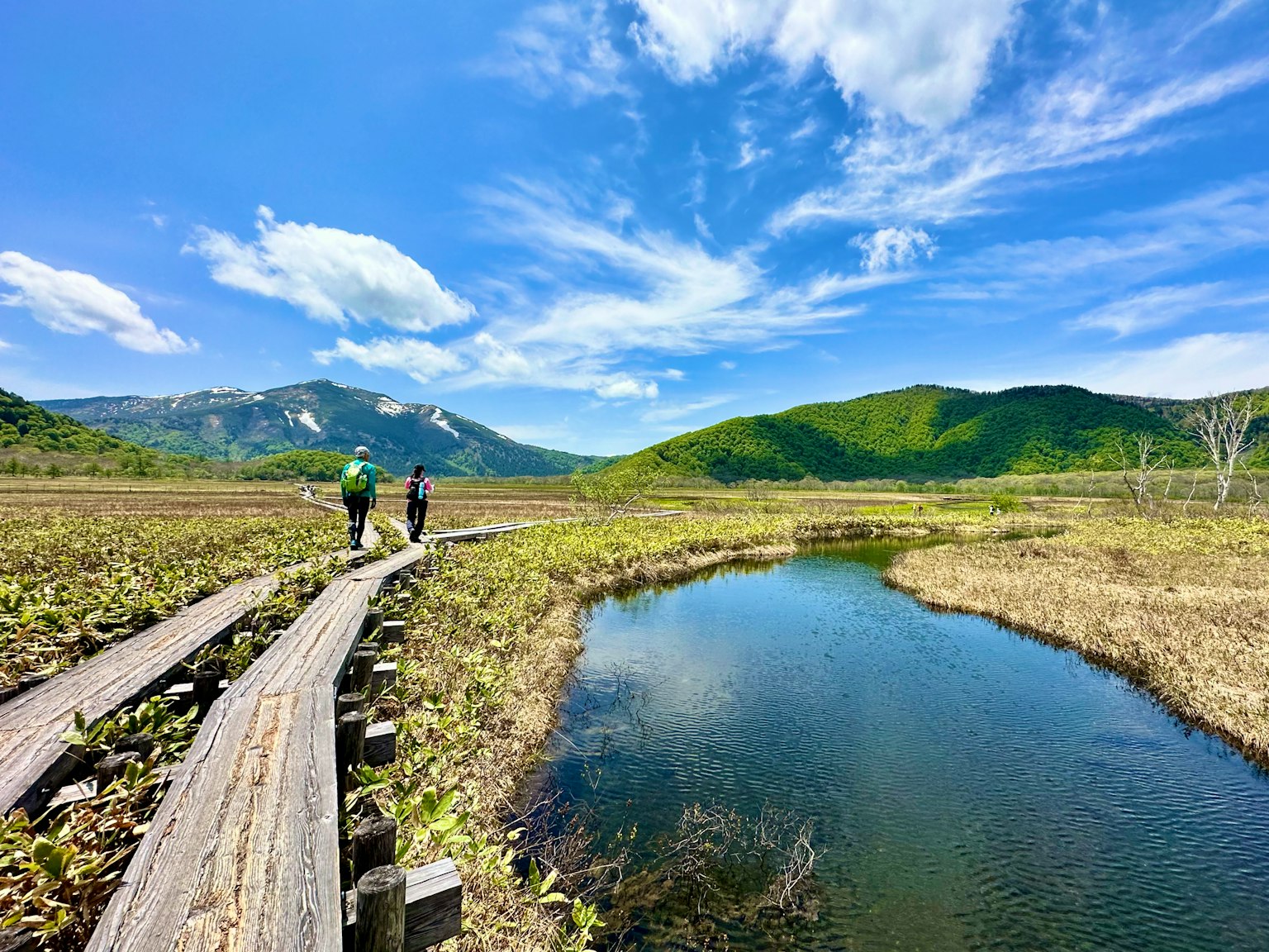 兩名登山健行者走在小池塘旁的木棧道上，背景是綠意盎然的山脈