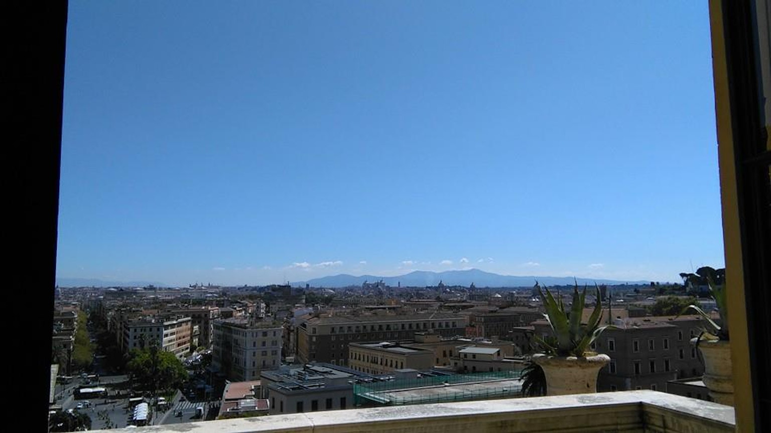 Panorama cittadino sotto un cielo blu chiaro con montagne lontane visibili attraverso una finestra