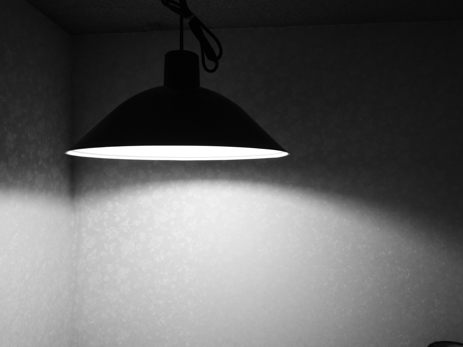 Una lampada a sospensione con paralume nero che illumina un muro in un interno semplice