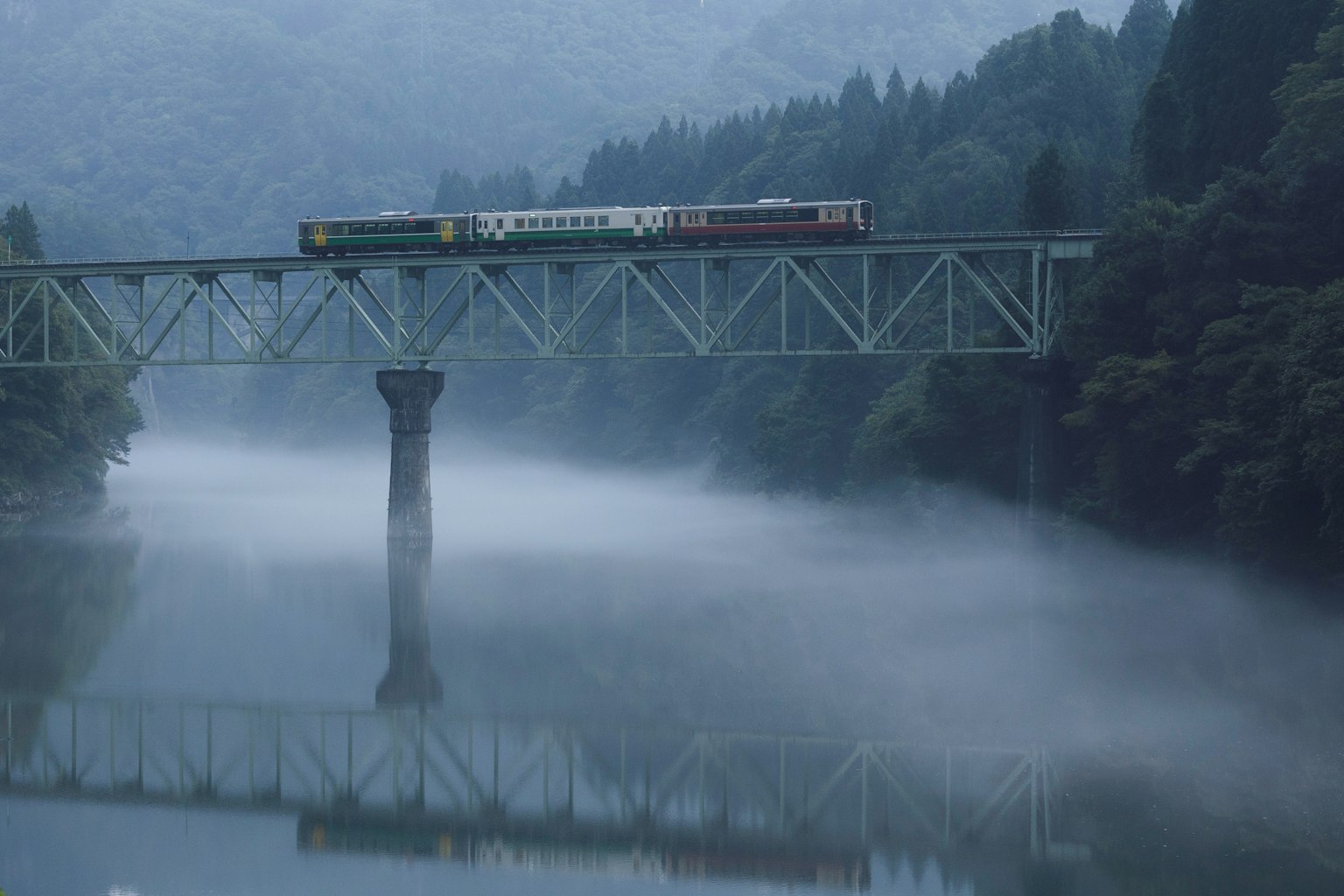 Train crossing a misty river bridge surrounded by mountains