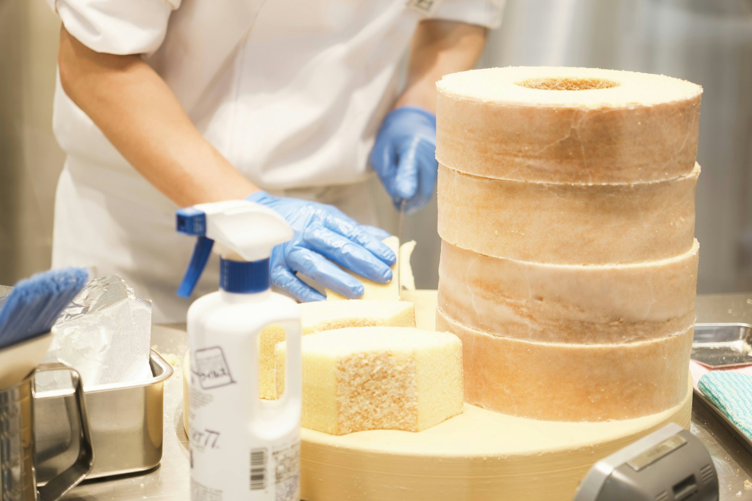 Un pasticcere con guanti blu che taglia una torta con torte impilate sullo sfondo