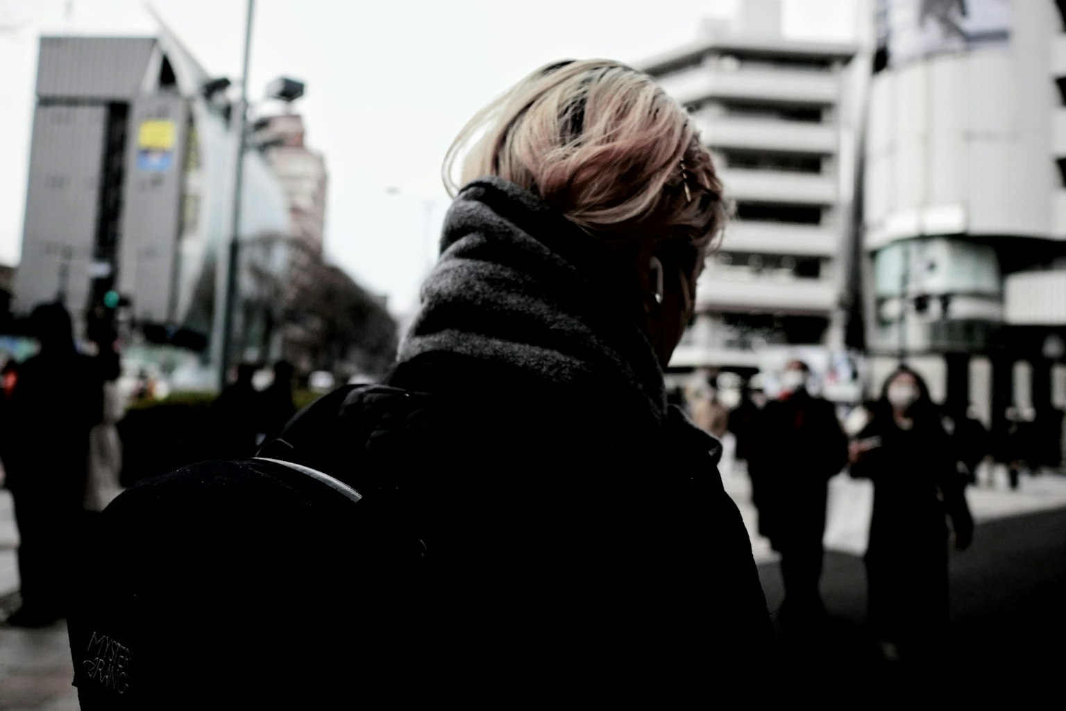 Person with light-colored hair seen from behind in a dark urban street setting with high-rise buildings
