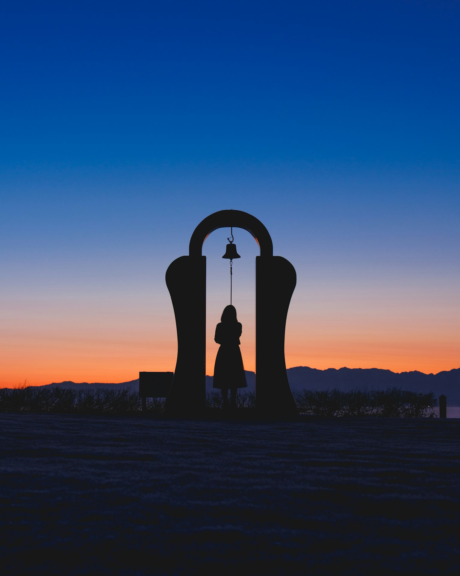 Silueta de una mujer junto a una campana al atardecer con montañas al fondo