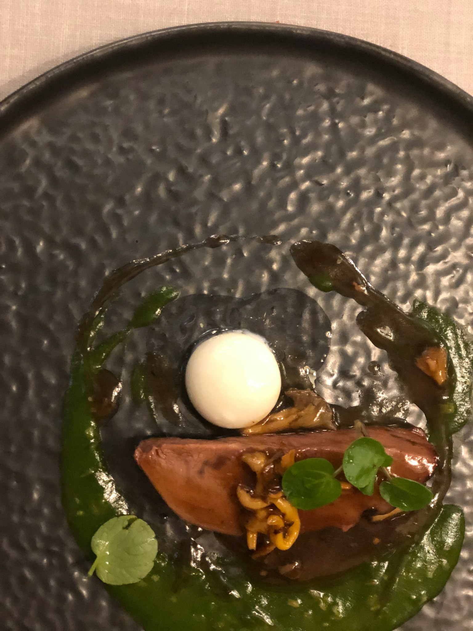 A plated dish on a black plate featuring a white spherical component and green sauce with nuts and herbs