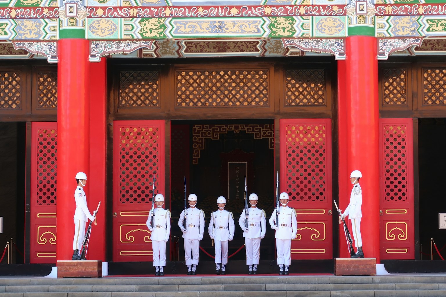 Lính gác trong đồng phục đứng thành hàng trước một tòa nhà truyền thống với các cột đỏ và trang trí tinh xảo