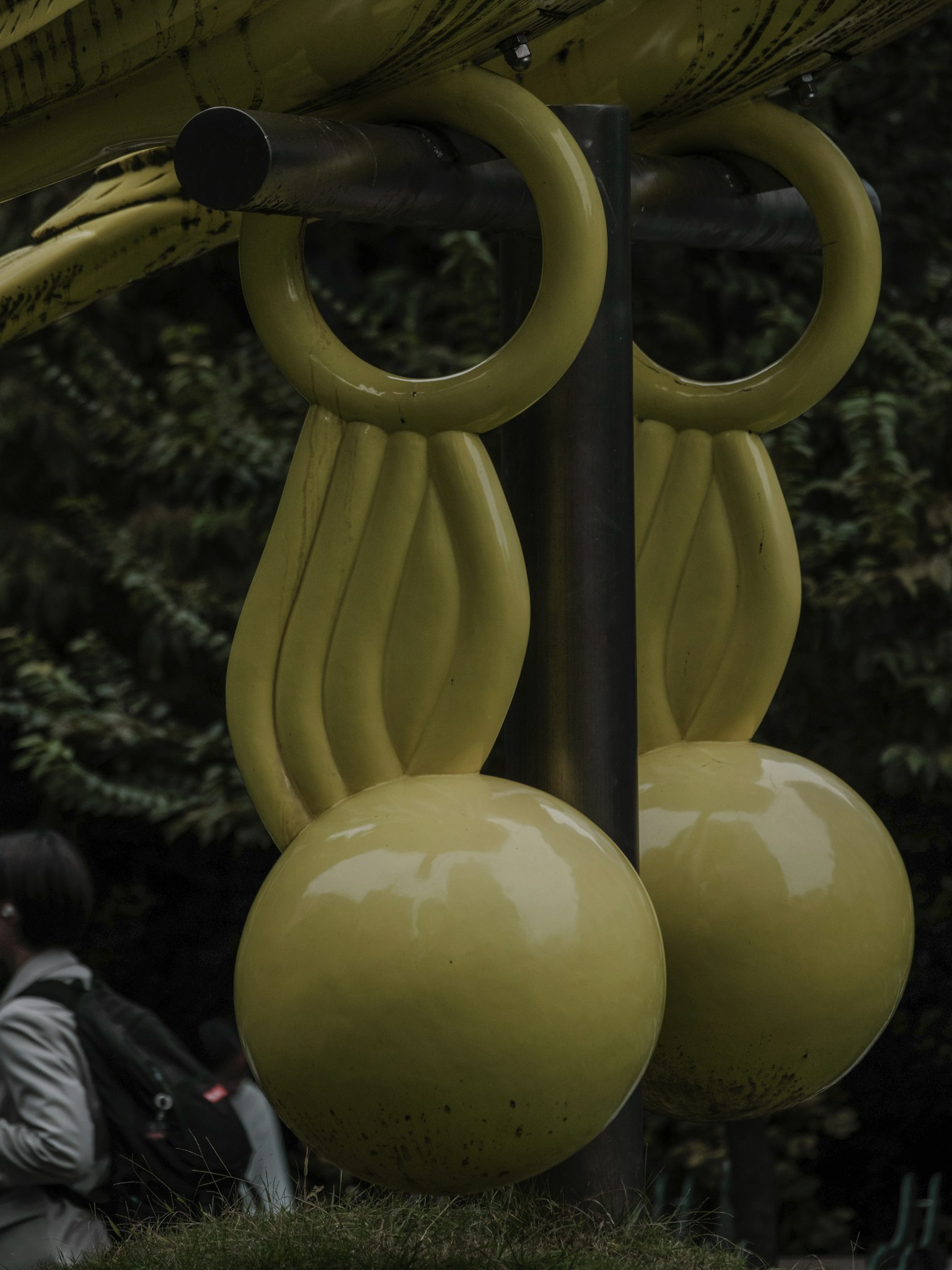 Part of a yellow playground structure in a park featuring a bird-like design with round balls and wing shapes