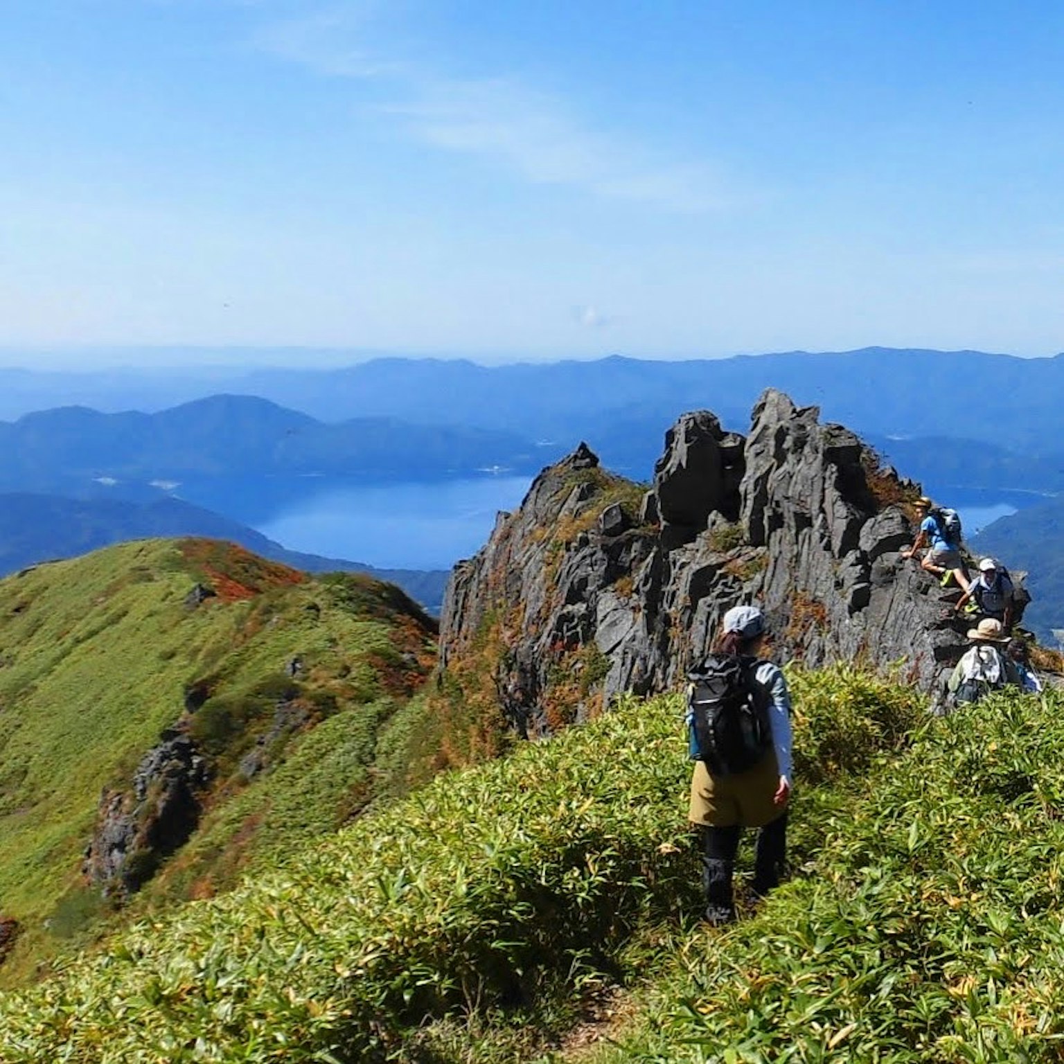 Menschen wandern auf einem grünen Bergrücken mit felsigen Gipfeln und einem entfernten See