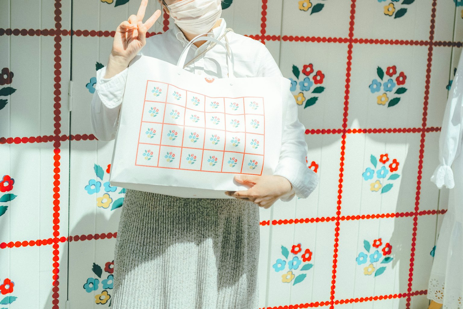 마스크를 쓴 여성이 꽃무늬 배경 앞에서 다채로운 토트백과 함께 포즈를 취하고 있다