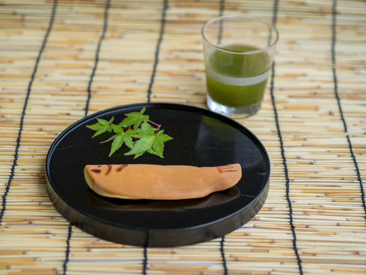 Un piatto nero con foglie verdi e un dolce giapponese accanto a un bicchiere di tè verde