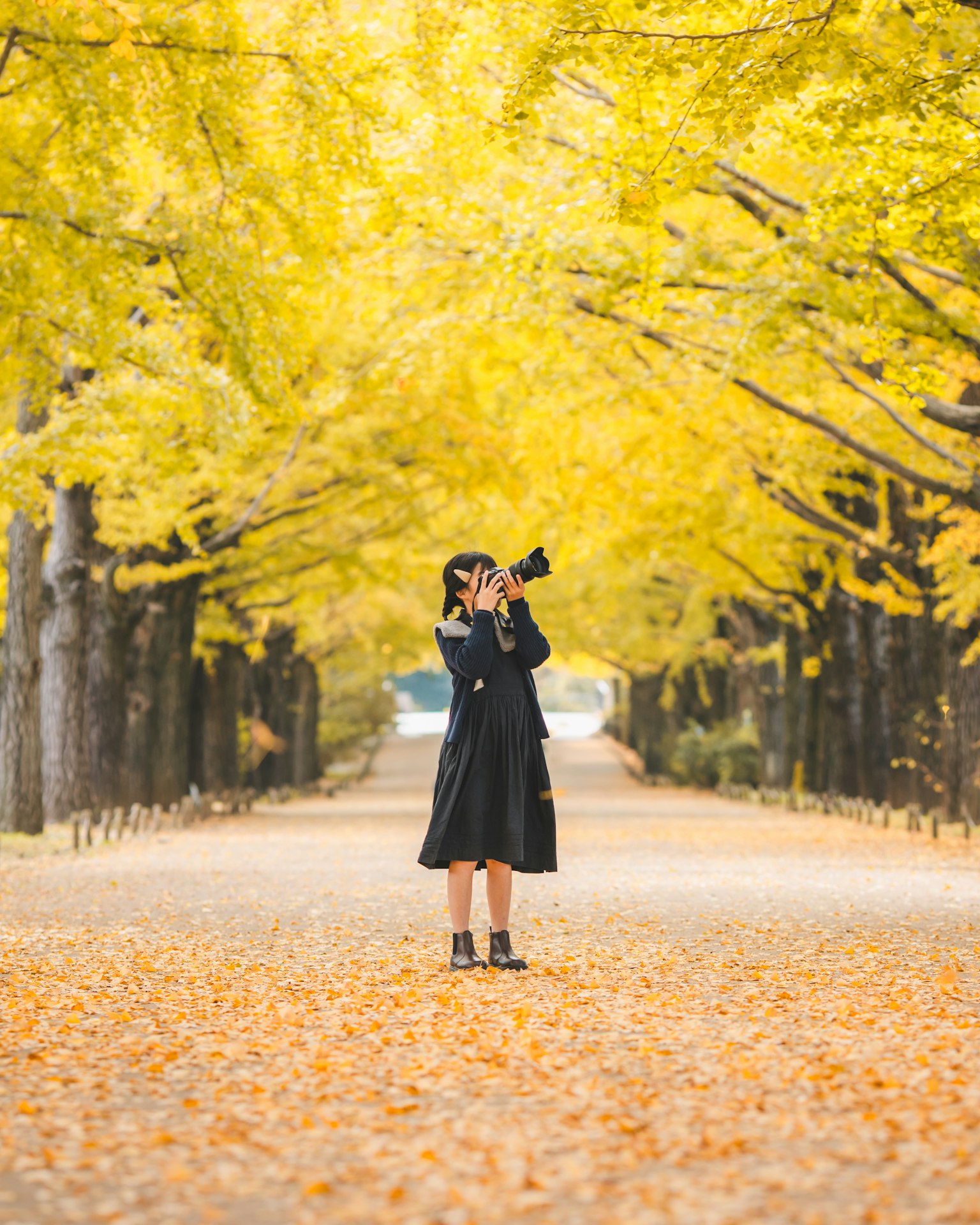 Una donna in piedi su un sentiero fiancheggiato da foglie gialle d'autunno che tiene una macchina fotografica