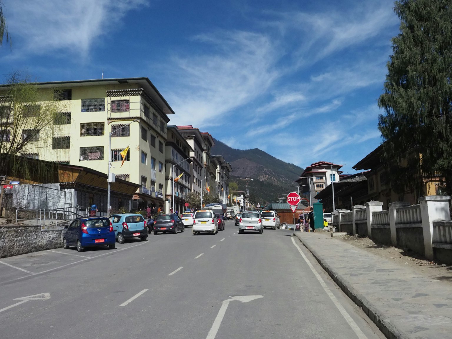Straßenszene in Bhutan mit blauem Himmel und Bergen im Hintergrund Gebäude und Autos entlang der Straße
