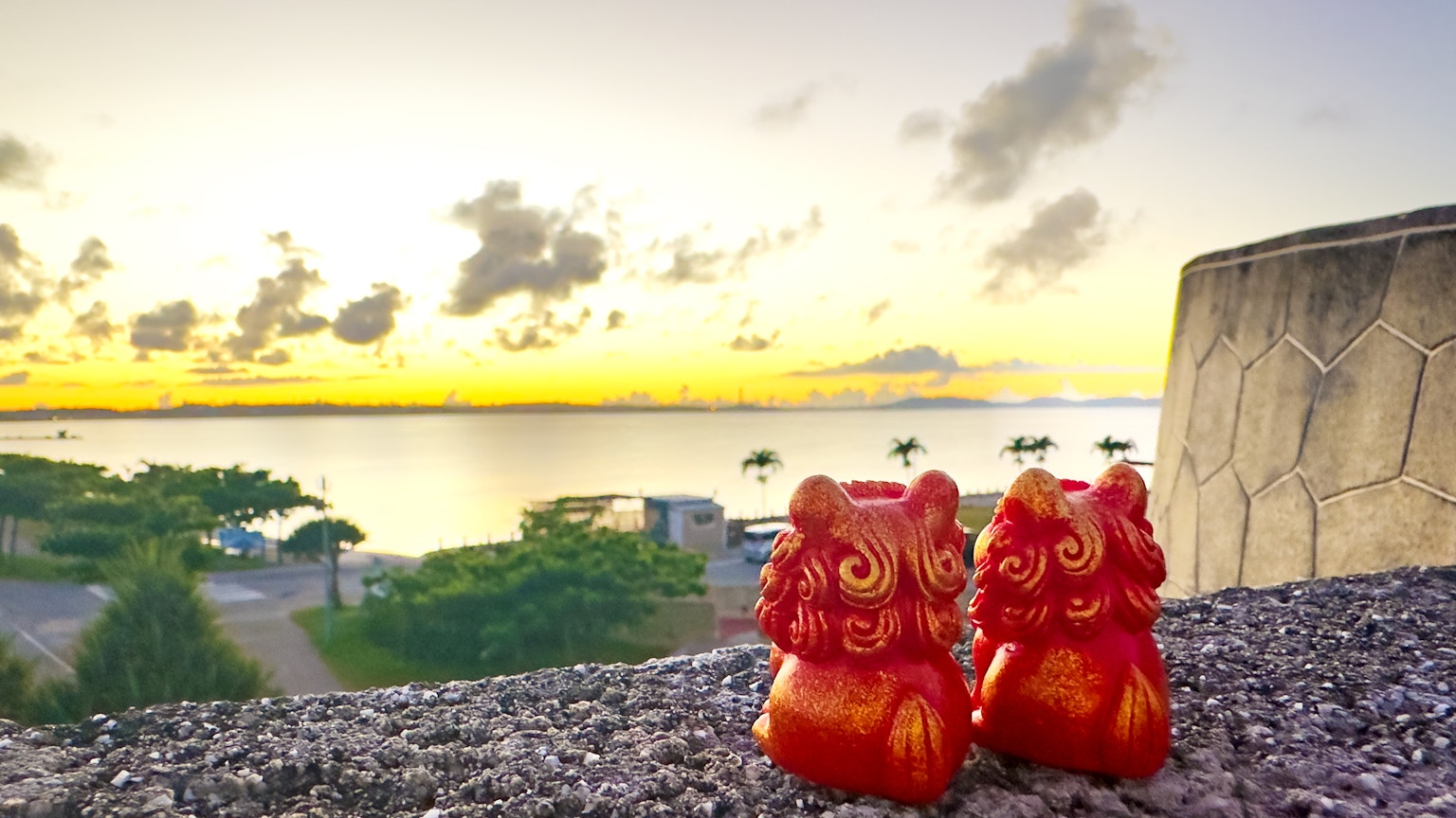 Dos estatuas rojas de Shisa en primer plano con un atardecer sobre el océano al fondo