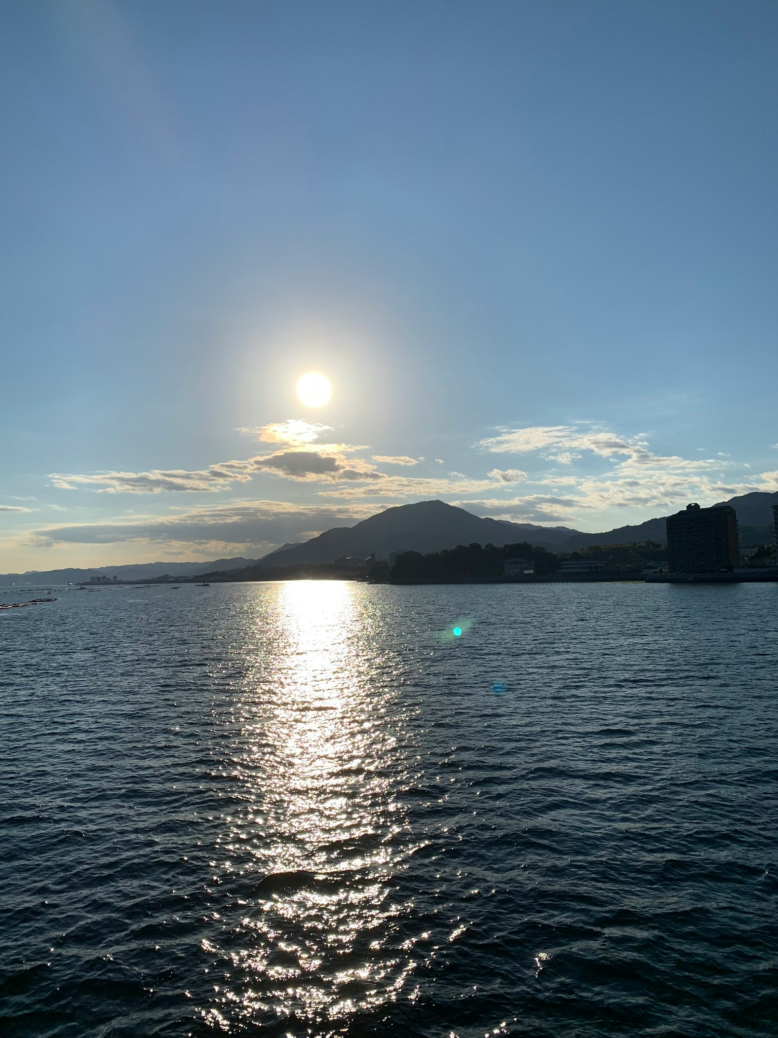Malersiche Aussicht auf das Meer mit der Sonne, die in einem blauen Himmel scheint, Silhouette von Bergen und schimmerndem Wasser