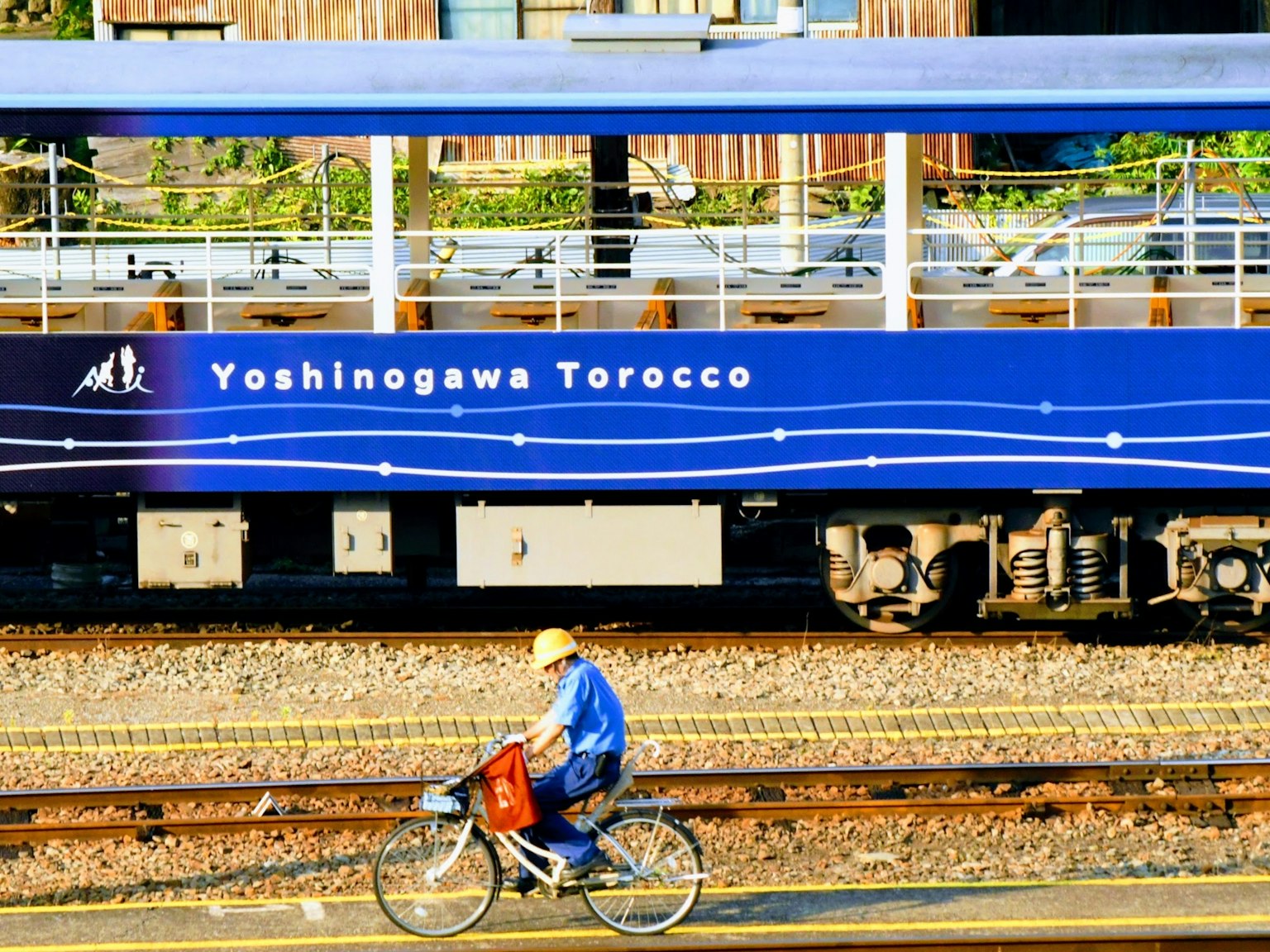 Adegan kereta biru dengan 'Yoshinogawa Torocco' tertulis di samping Seseorang yang mengendarai sepeda melintas