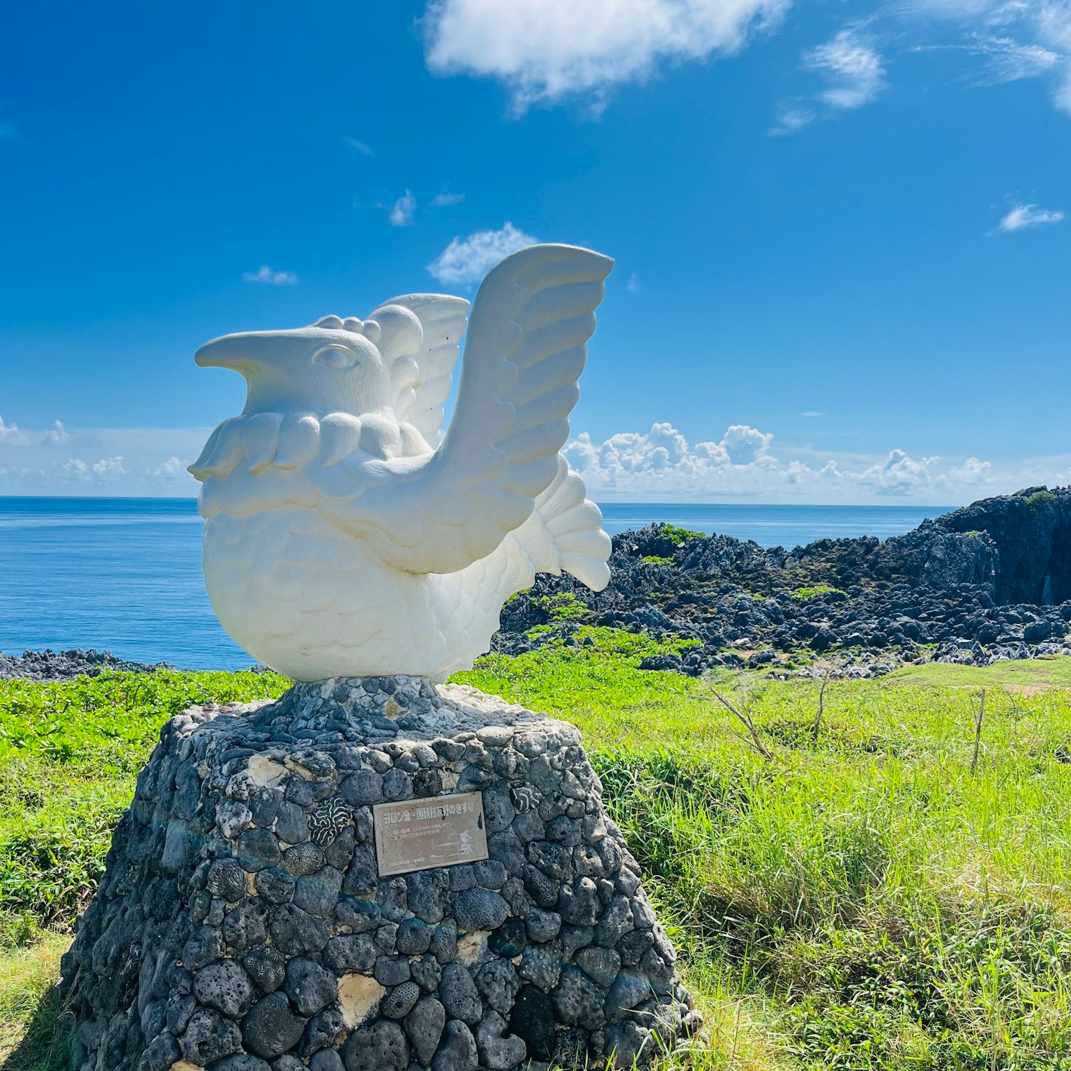 Escultura blanca de un pájaro contra el mar azul y el cielo con costa rocosa