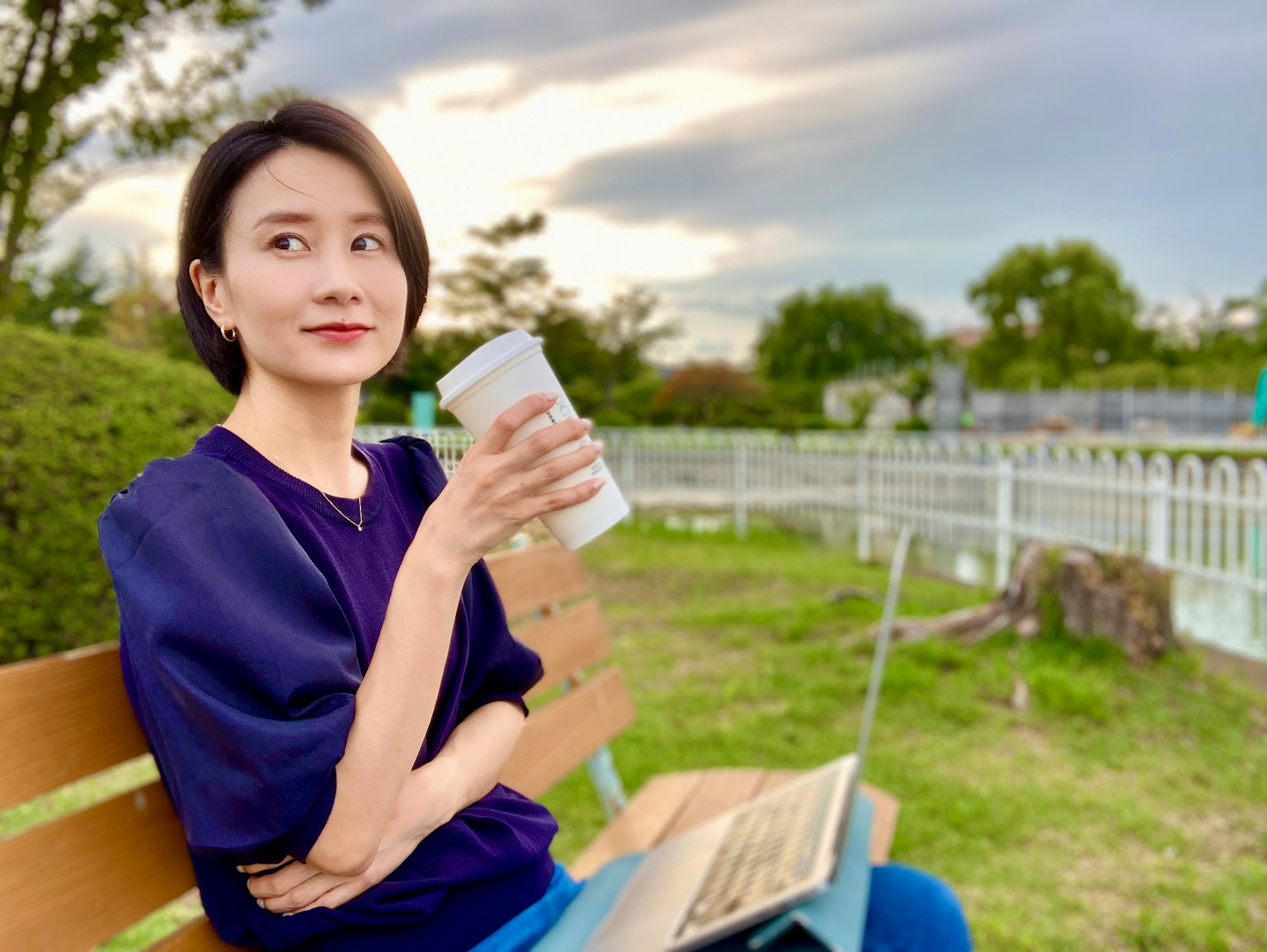 ผู้หญิงนั่งบนม้านั่งในสวนถือแก้วกาแฟอยู่หน้าคอมพิวเตอร์โน้ตบุ๊ก