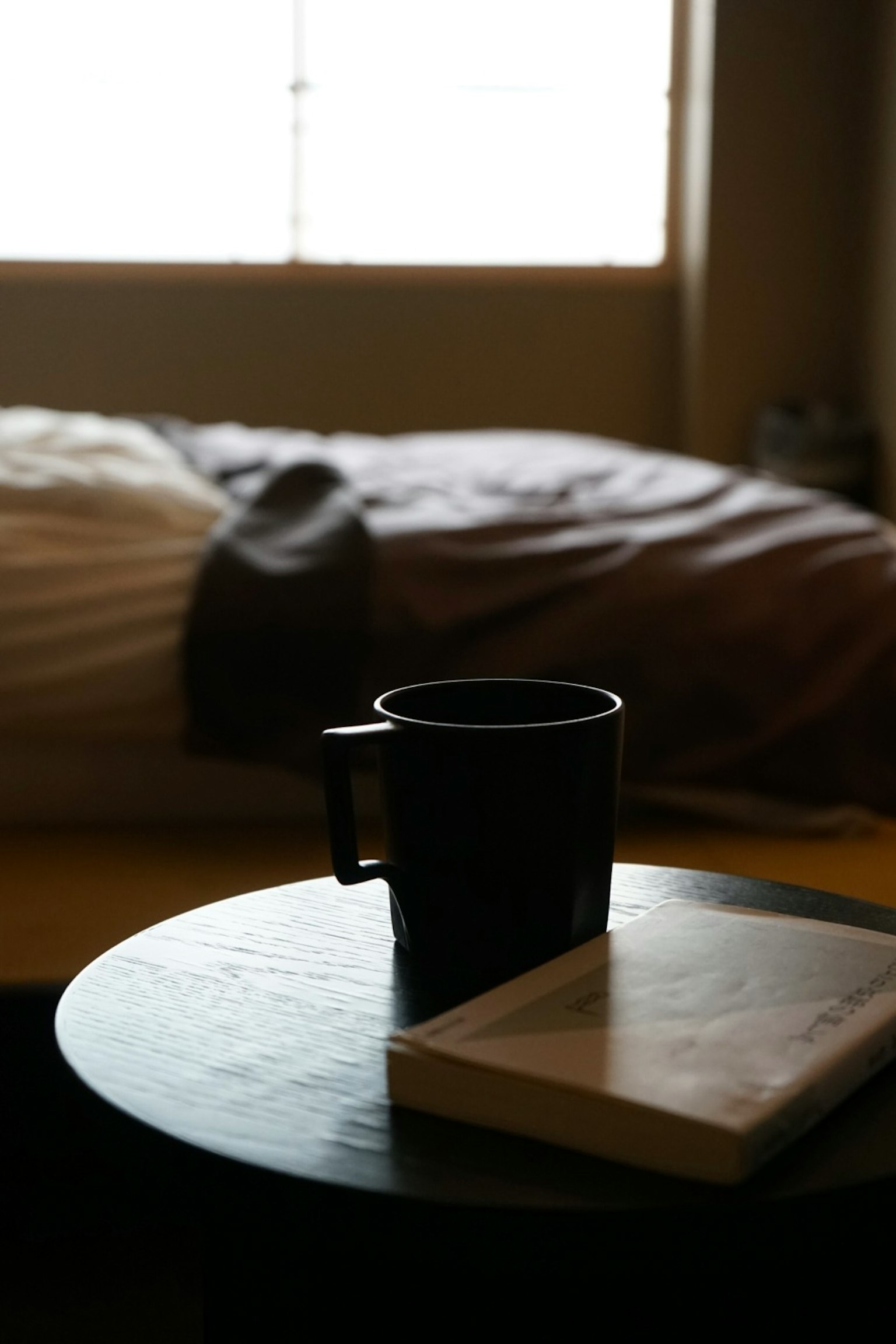 ベッドと本が置かれたテーブルにコーヒーカップがあるシンプルな室内の風景