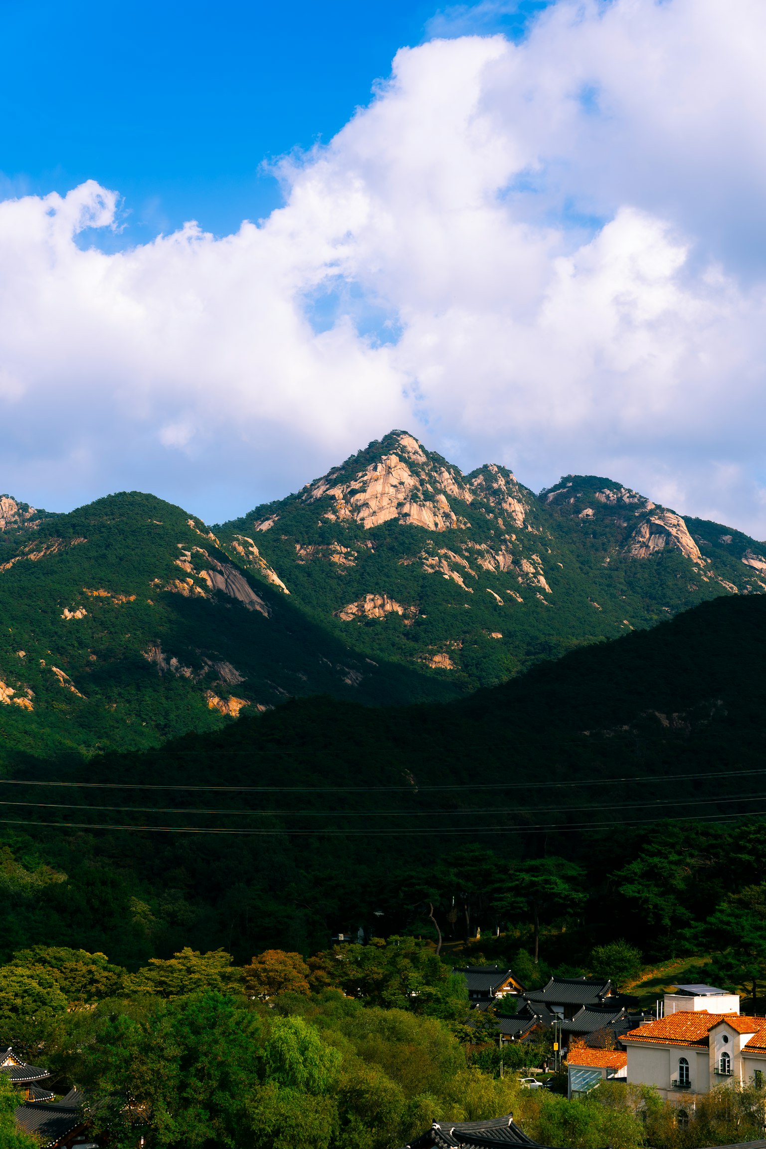 青空と白い雲の下にそびえる山々の風景 緑豊かな木々と住宅が見える