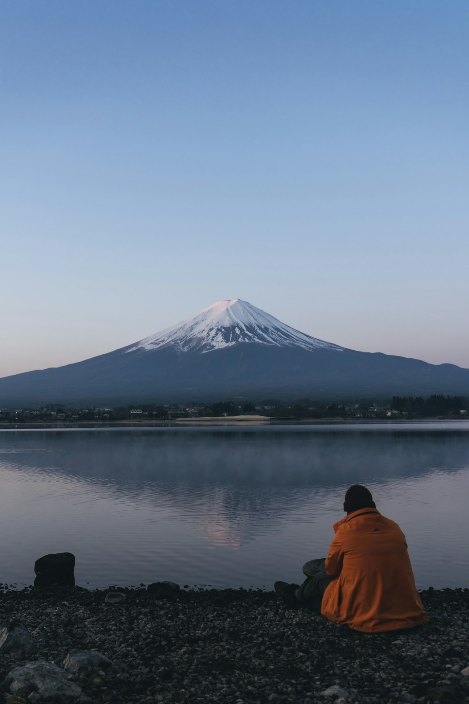 A person in an orange coat sitting by a calm lake with Mount Fuji in the background