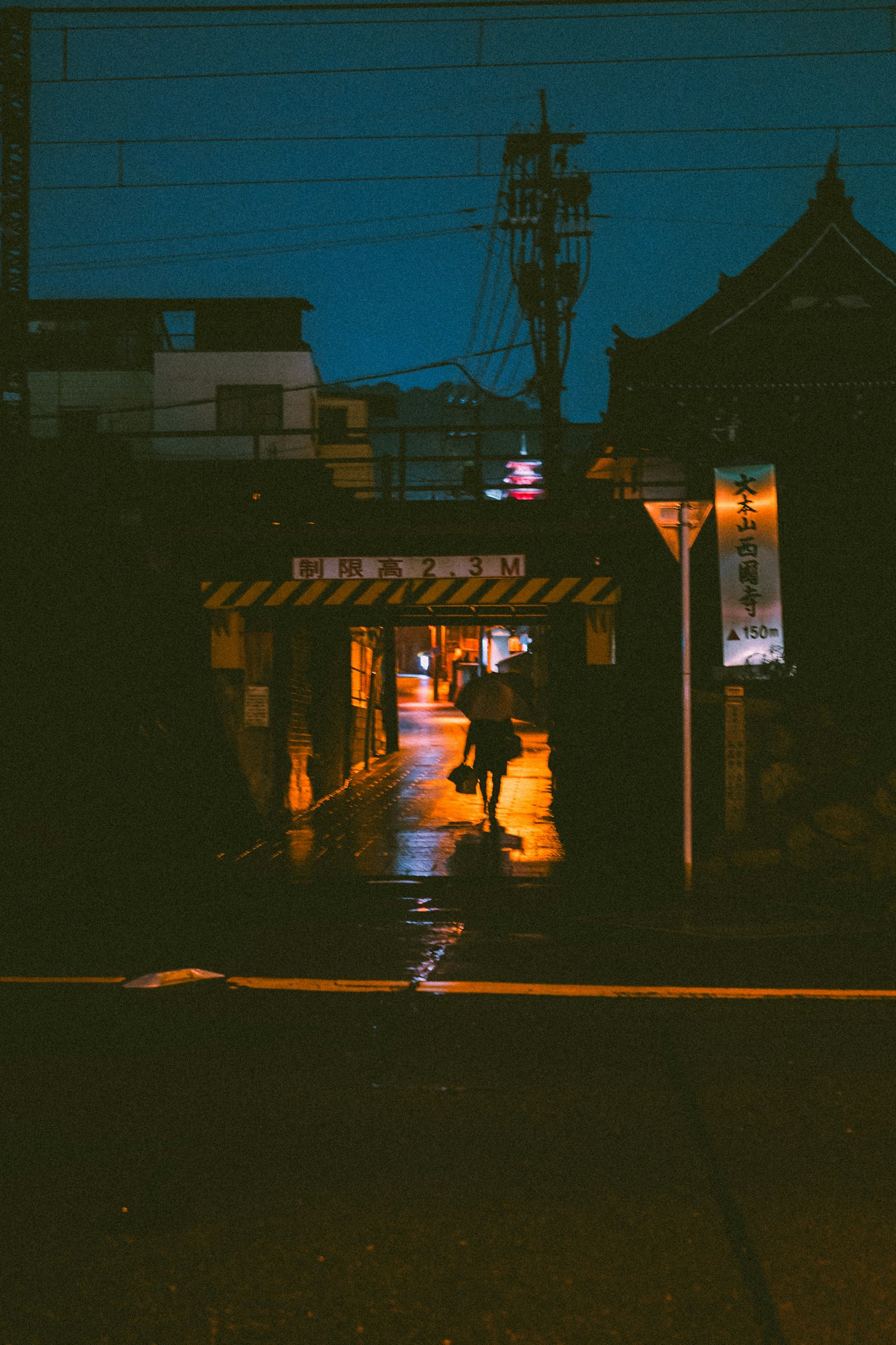 一個人在雨中濕透的小巷裡行走，夜晚被橙色路燈照亮