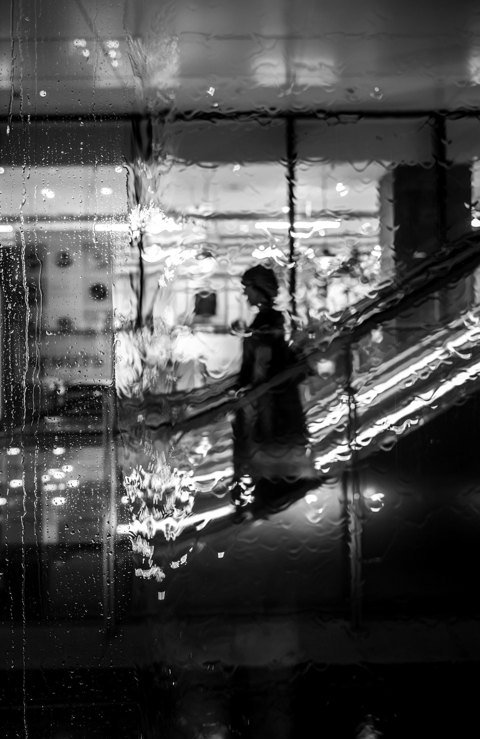 黒と白の写真で、階段を下りる人が水滴のあるガラス越しに映っている