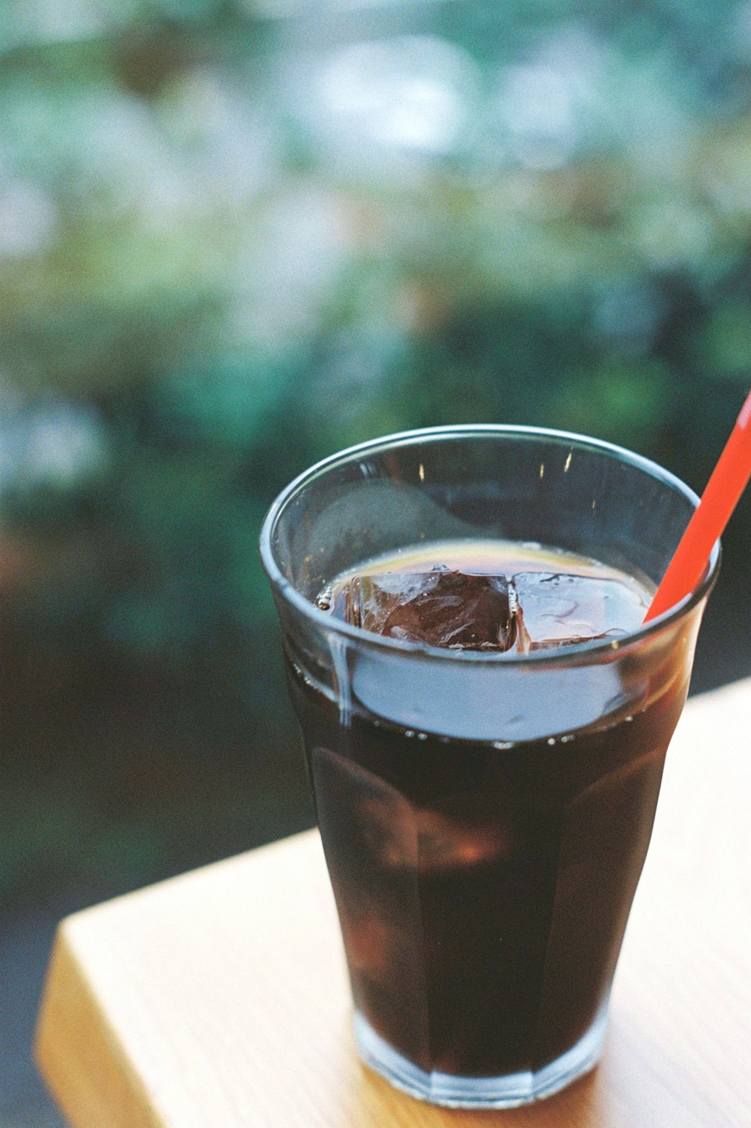 Secangkir kopi es dalam gelas transparan dengan es batu dan sedotan merah, dikelilingi latar belakang hijau yang kabur