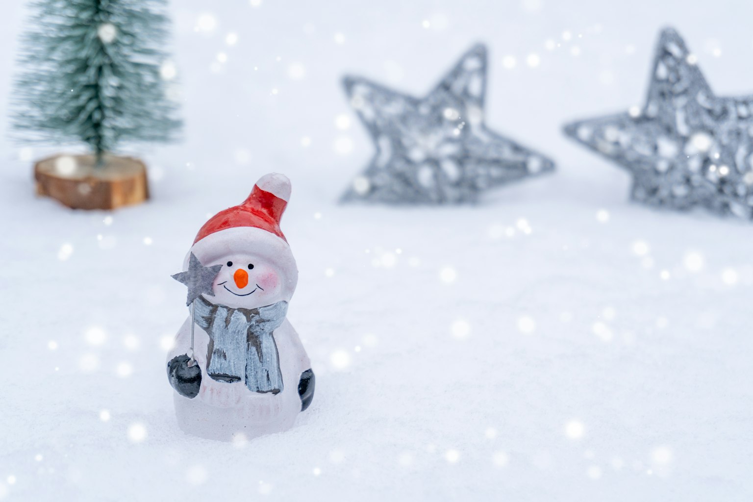 Schneemannfigur auf Schnee mit rotem Hut und blauer Schal kleiner Weihnachtsbaum und silberne Sterne im Hintergrund