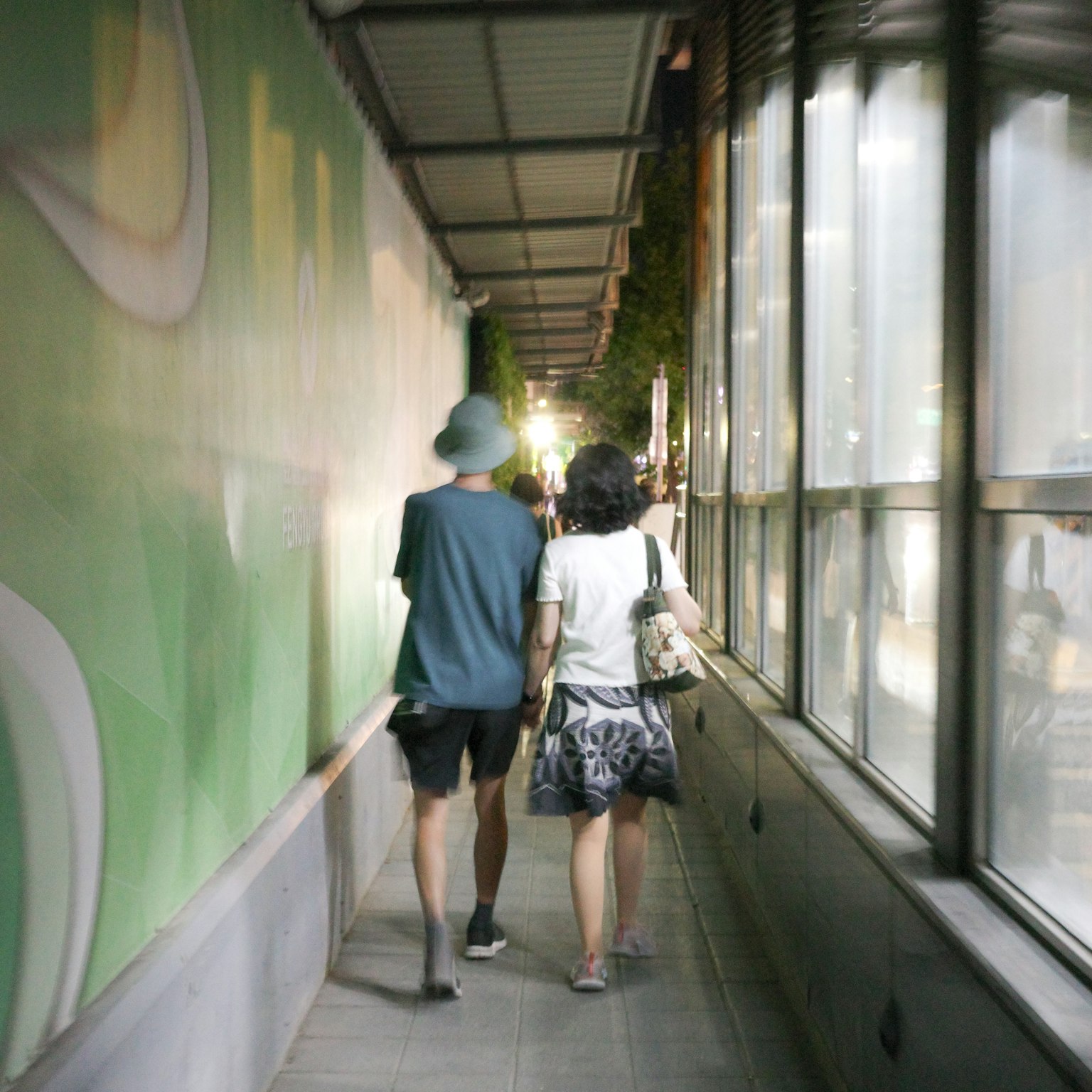 Pareja caminando por una acera estrecha con una pared verde y ventanas de vidrio