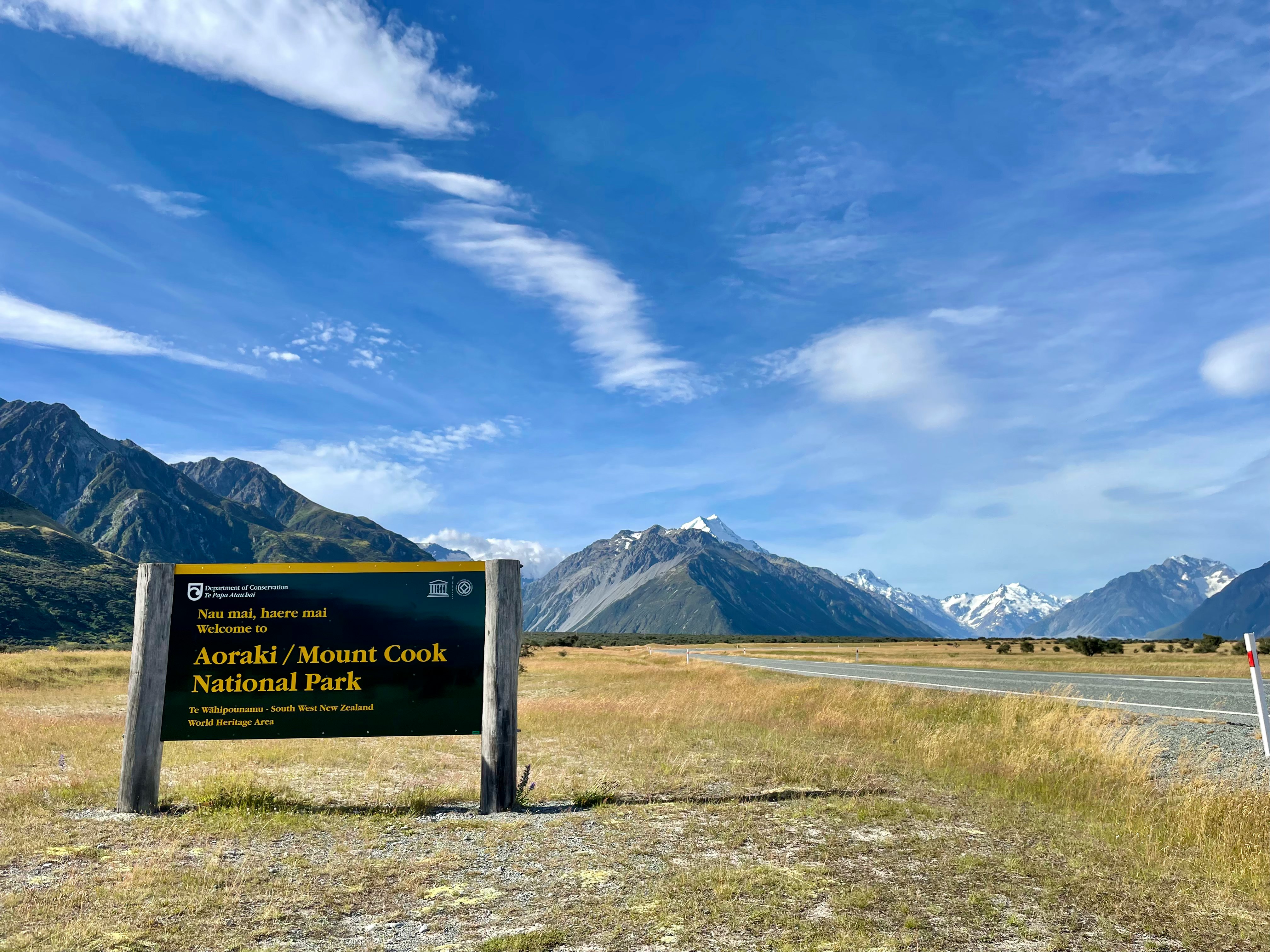 Schild „Aoraki/Mount Cook National Park“ an einem Weg in einer Wiese mit Berggipfeln