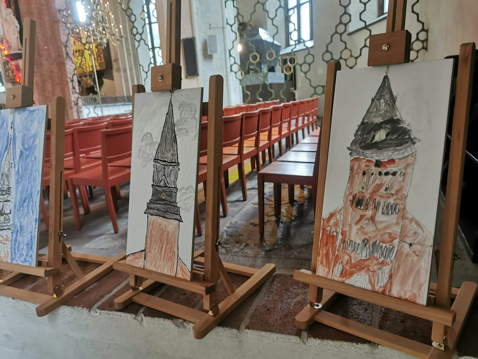 Tres dibujos de niños expuestos en caballetes dentro de una iglesia