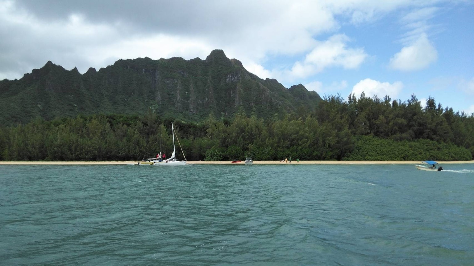 ทิวทัศน์ที่สวยงามมีเรือใบอยู่ใกล้ชายหาดทรายและภูเขาเขียวชอุ่มในพื้นหลัง