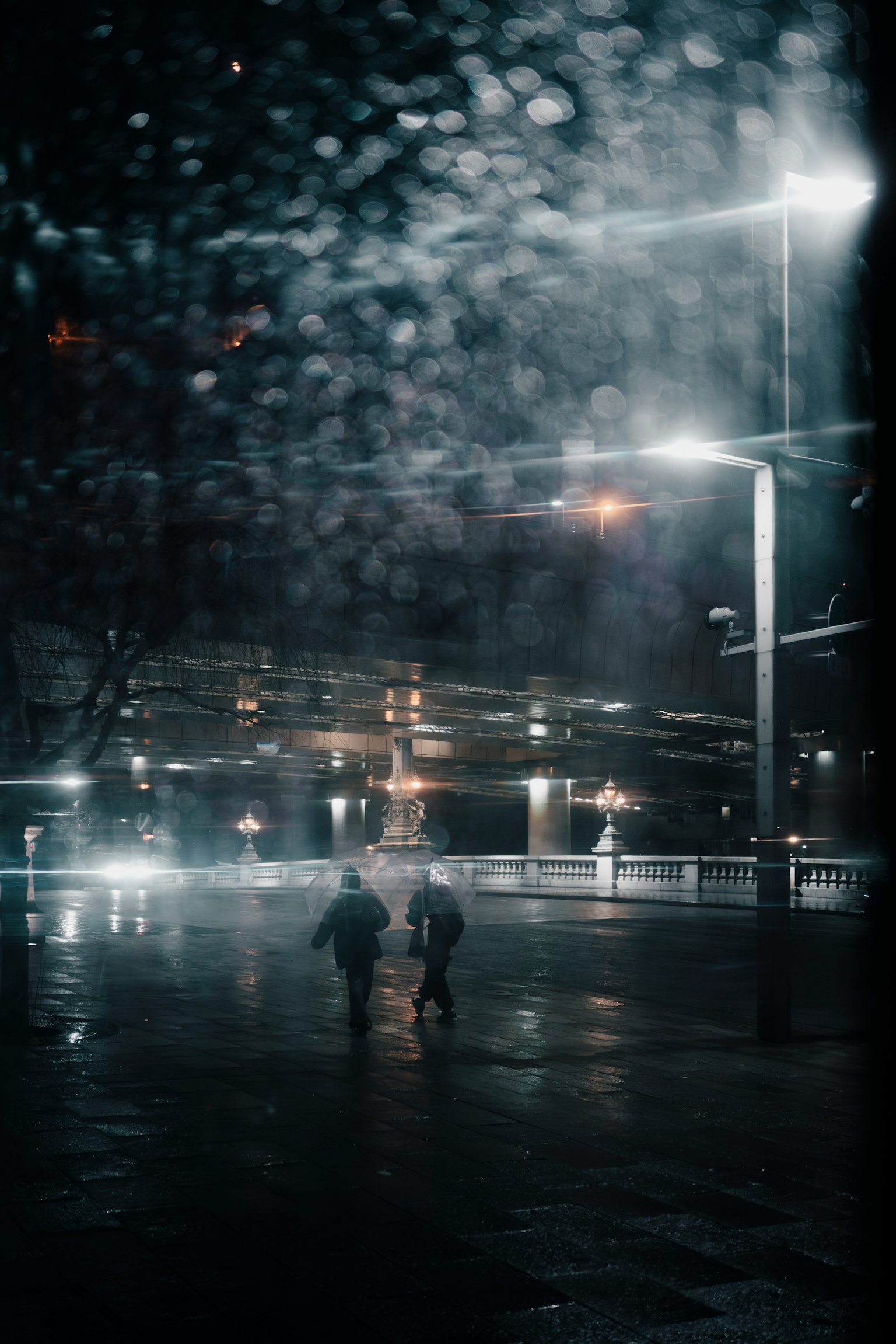 Dos figuras caminando bajo la lluvia por la noche con paraguas iluminados por farolas y reflejos en el pavimento mojado