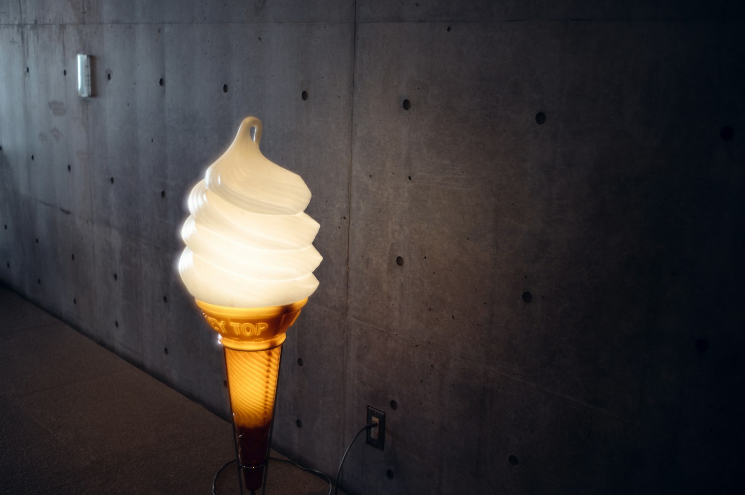 A lamp shaped like soft serve ice cream stands in front of a concrete wall