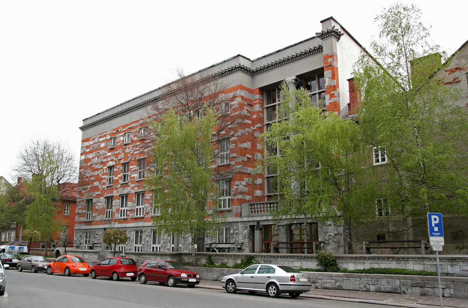 Edificio moderno con esterno arancione e grigio, circondato da alberi verdi e parcheggio, paesaggio urbano