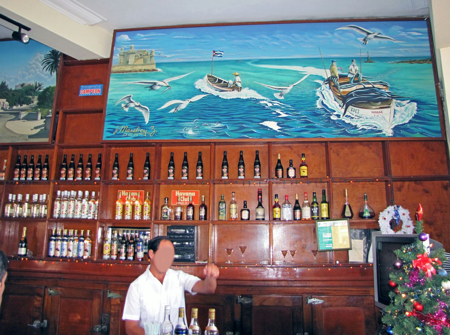 Interior de un bar con un mural de paisaje marino y un mostrador lleno de botellas