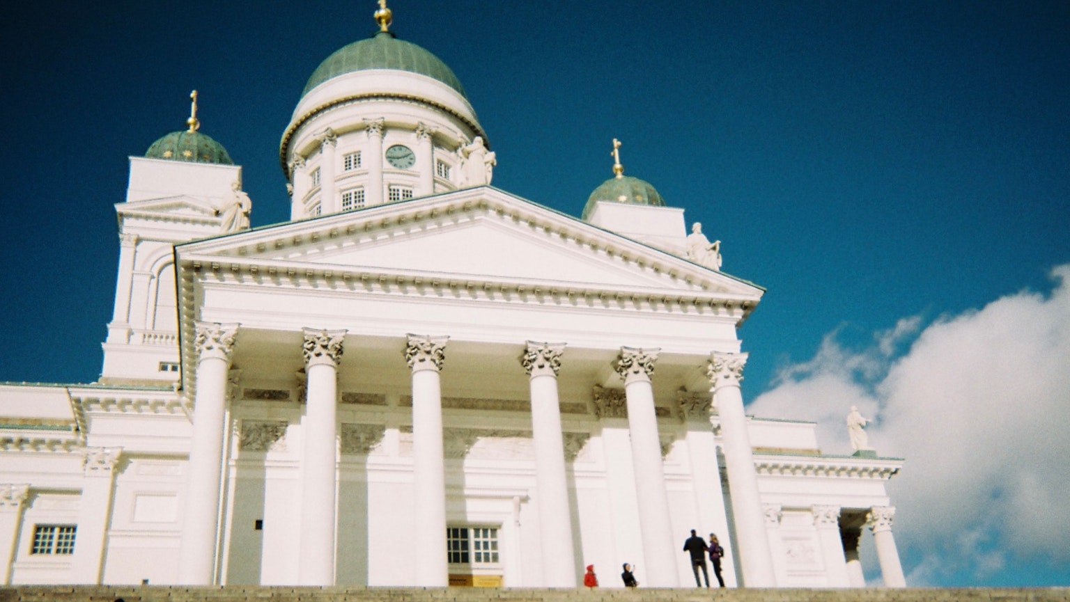Catedral de Helsinki con exterior blanco y cúpulas verdes bajo un cielo azul con personas