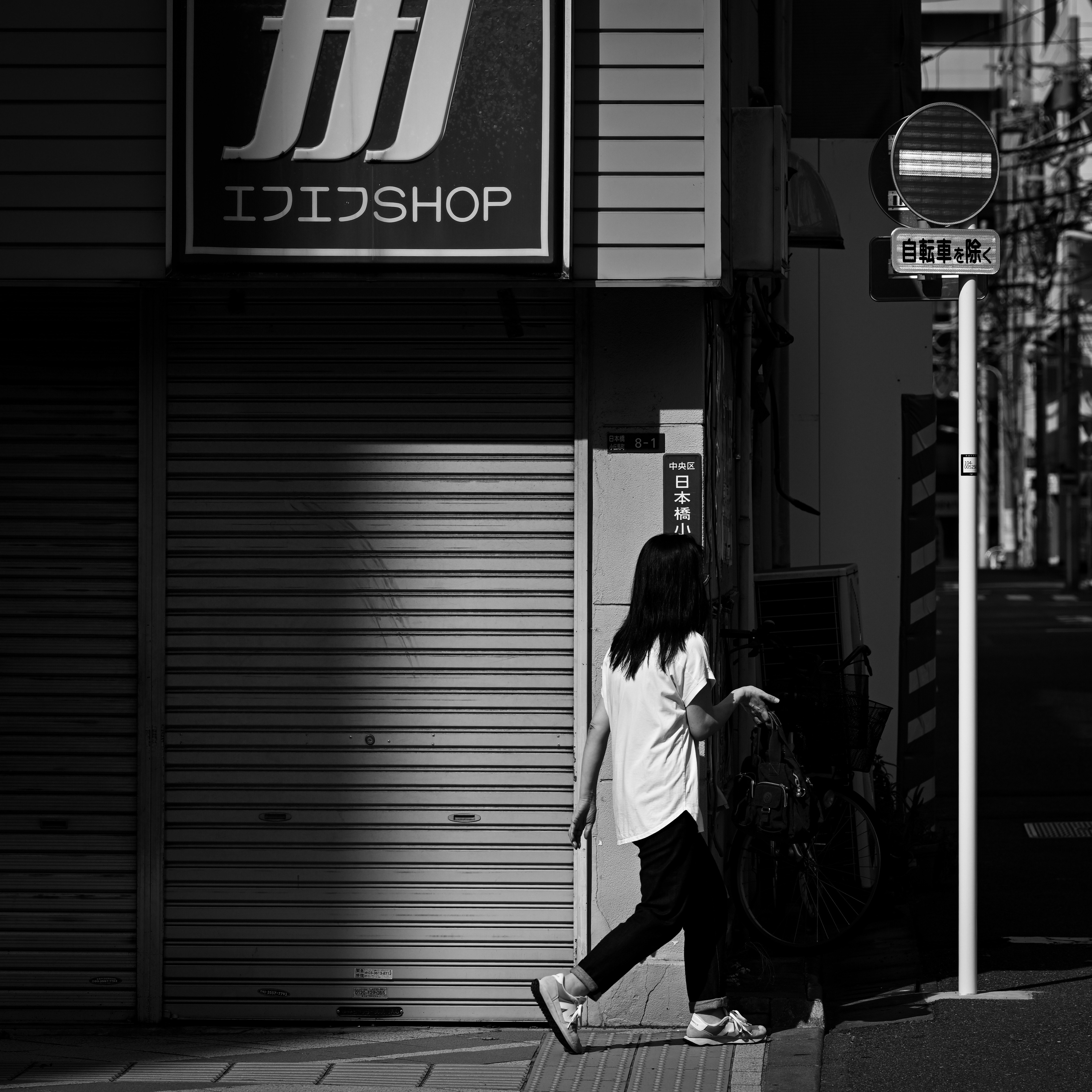 曲がり角の路地で自転車の横に立つ女性と光と影のモノクロ東京の商店街の軒先