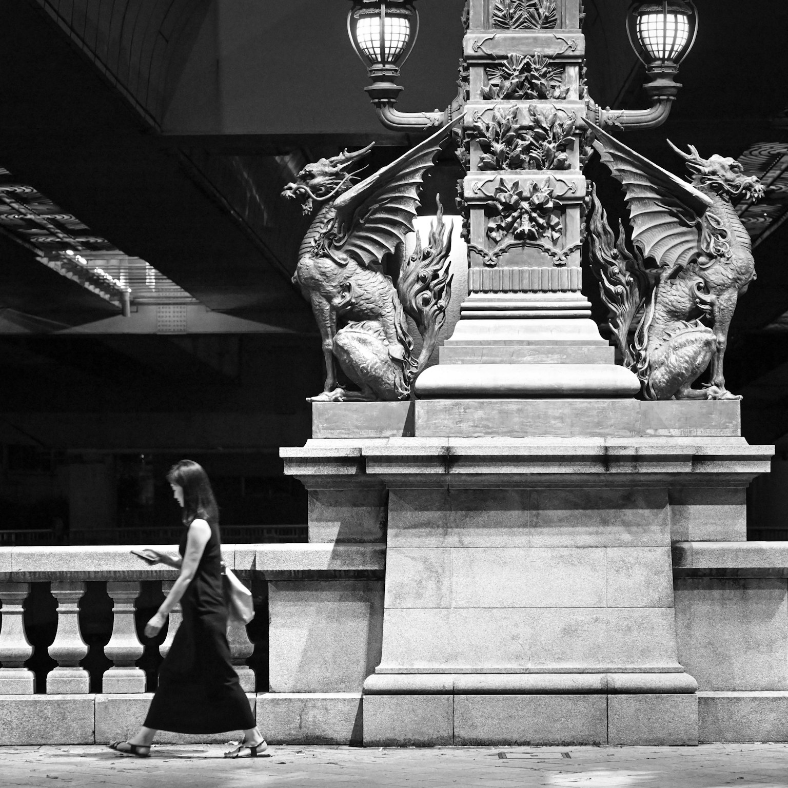 Una mujer caminando cerca de un pilar de puente en blanco y negro con esculturas de dragones