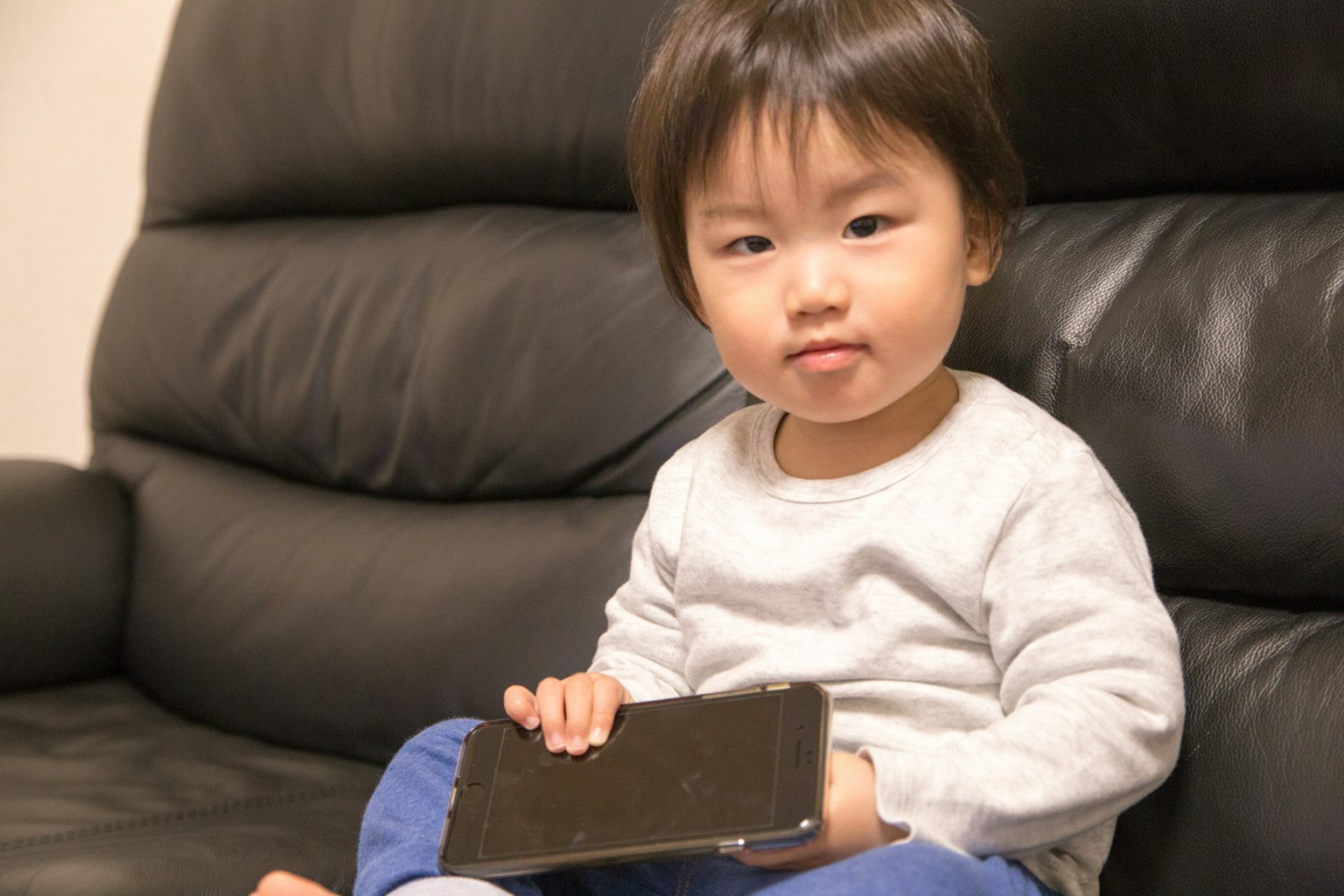 Ein kleines Kind sitzt auf einem schwarzen Sofa und hält ein Smartphone