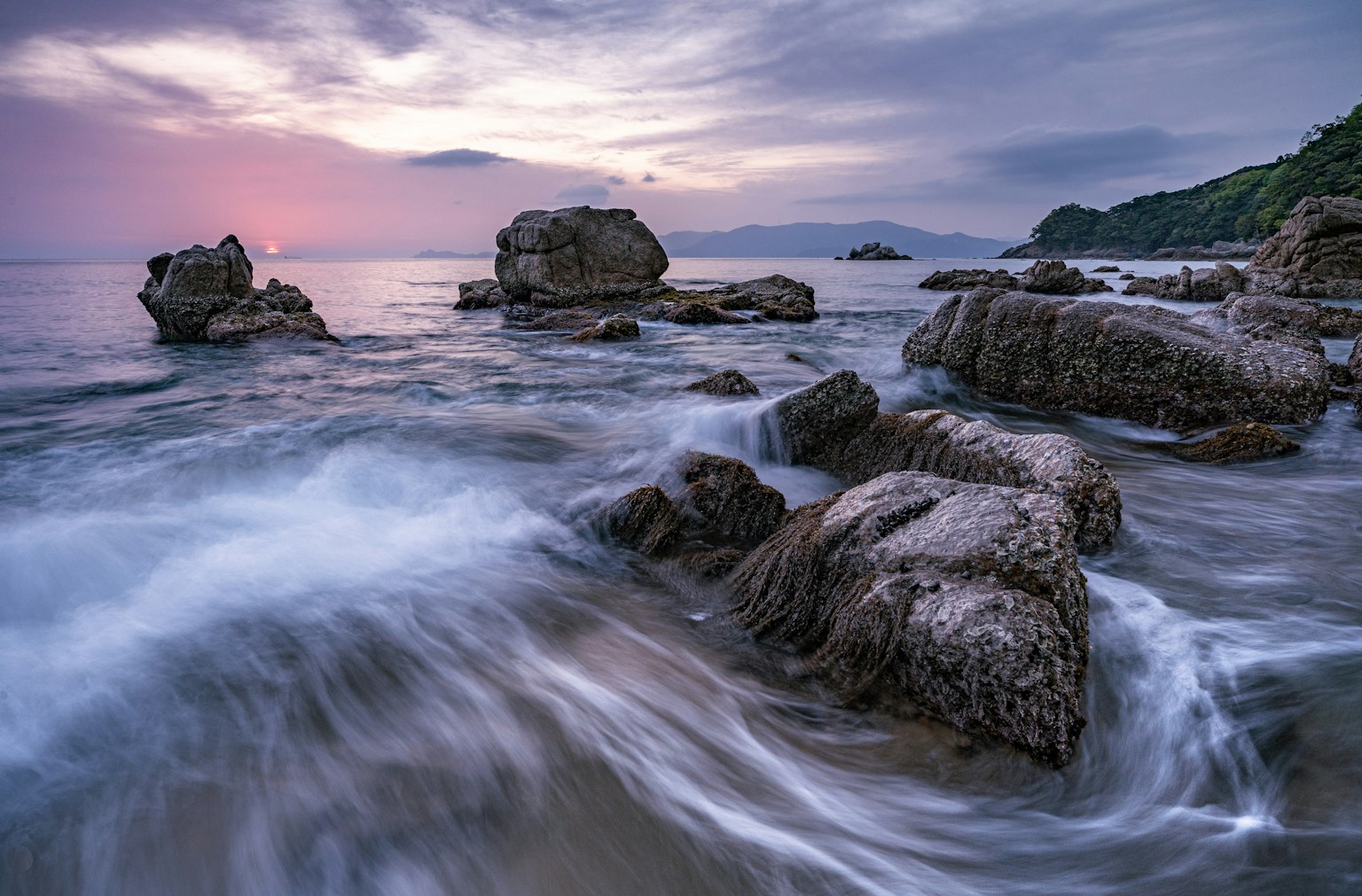 Matahari terbenam di lautan dengan garis pantai berbatu dan ombak lembut